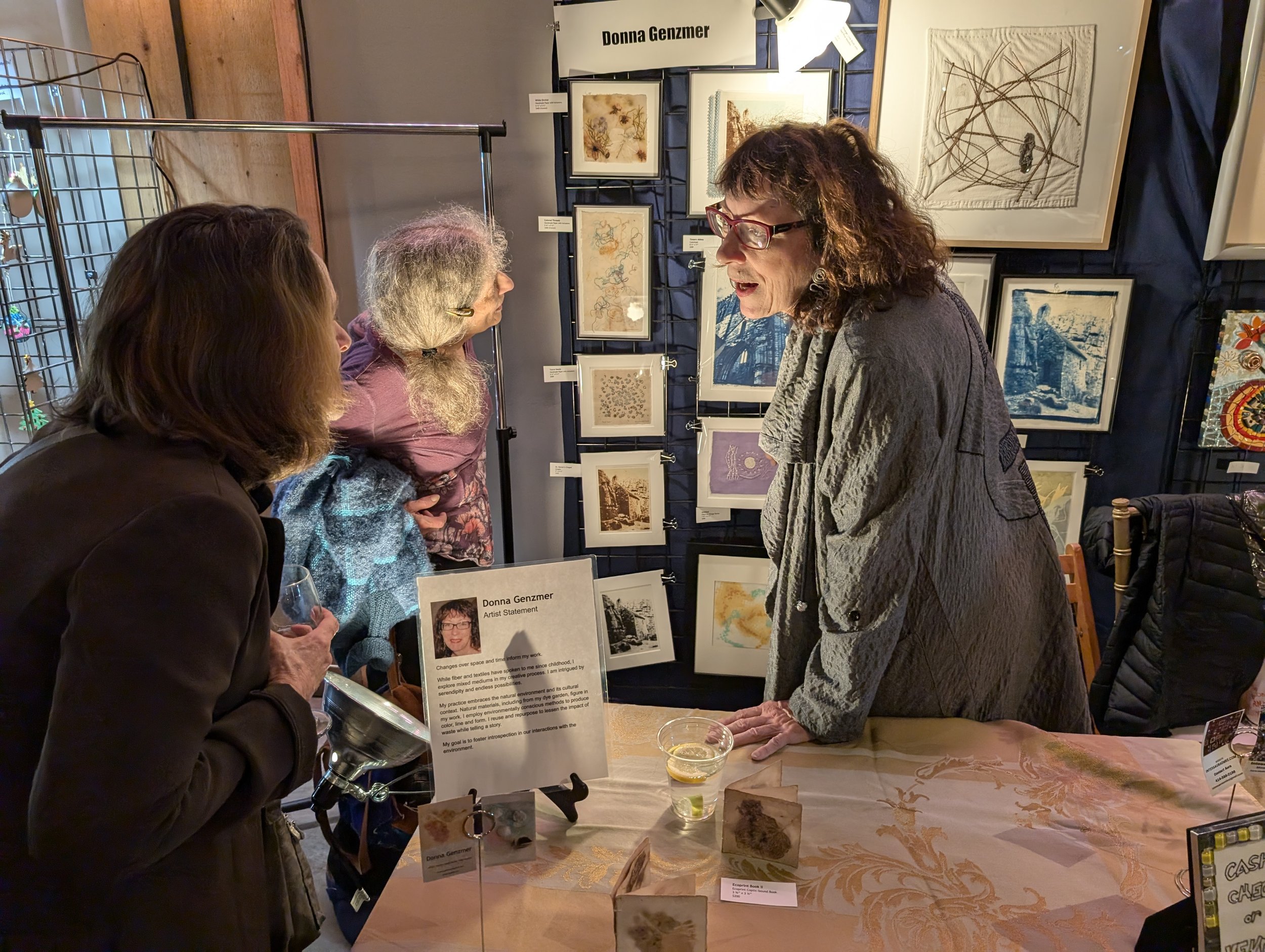Three women are engaged in conversation at an art exhibition. One woman behind the table is speaking to two women in front of her, with framed artwork displayed on the wall behind them. The table has a sign with information about Donna Genzmer, inclu