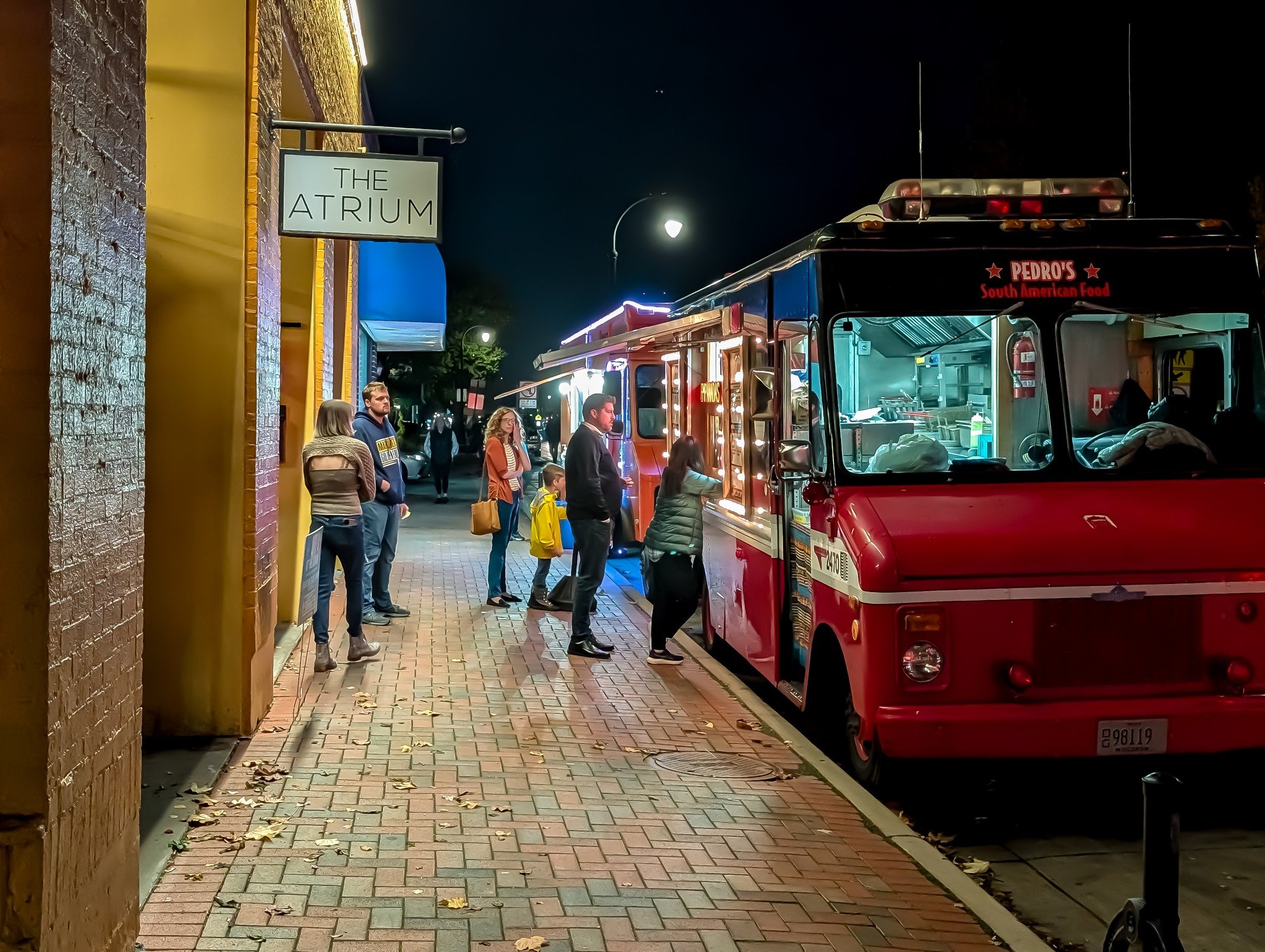 Night scene on a city sidewalk with people ordering from a red food truck labeled 'Pedro's South American Food' near a building with a sign for 'The Atrium'. The food truck is illuminated and has an open window where customers are served. A few leave