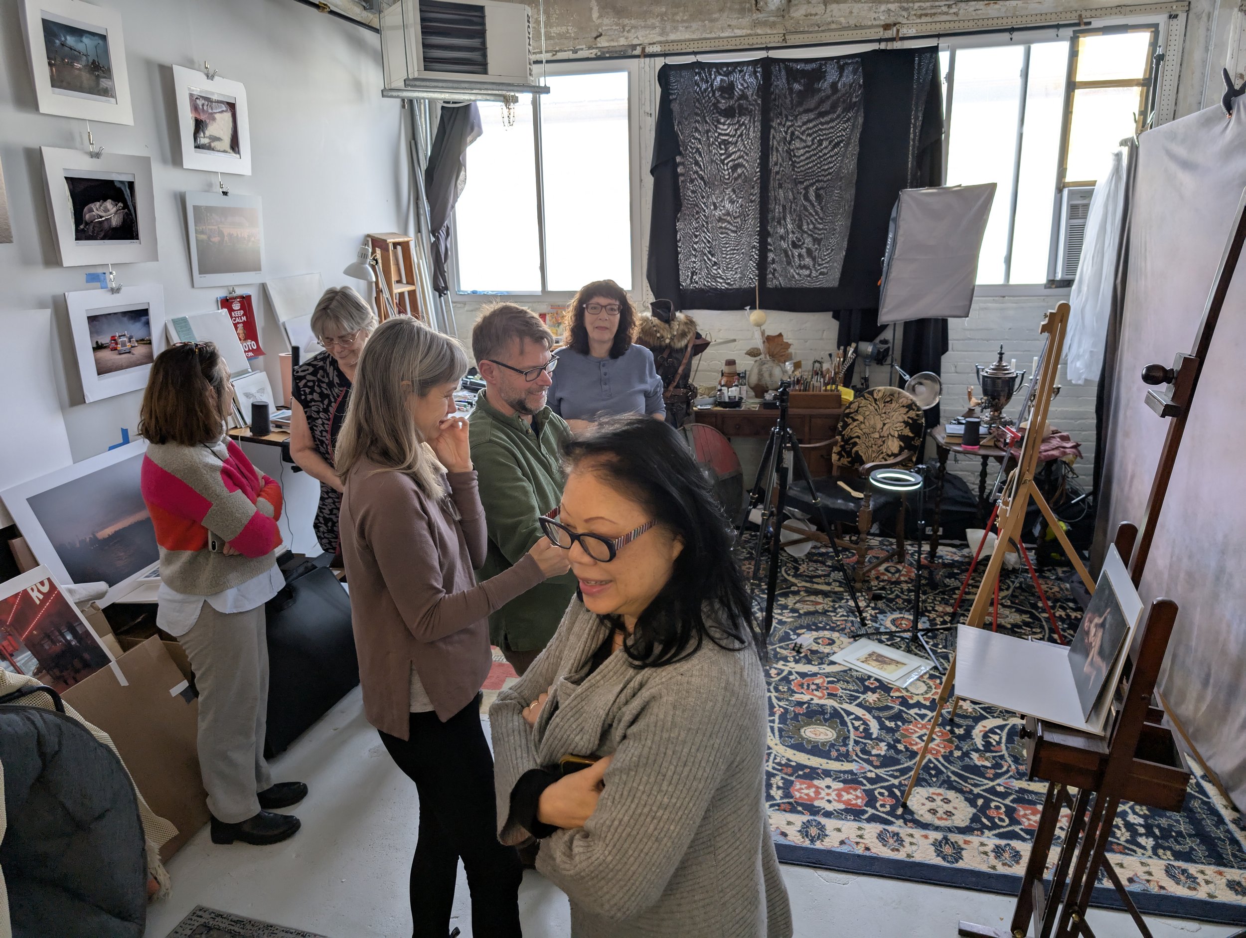 A group of people inside an art studio or gallery, engaging in conversation and observing art pieces displayed on the walls and easels.