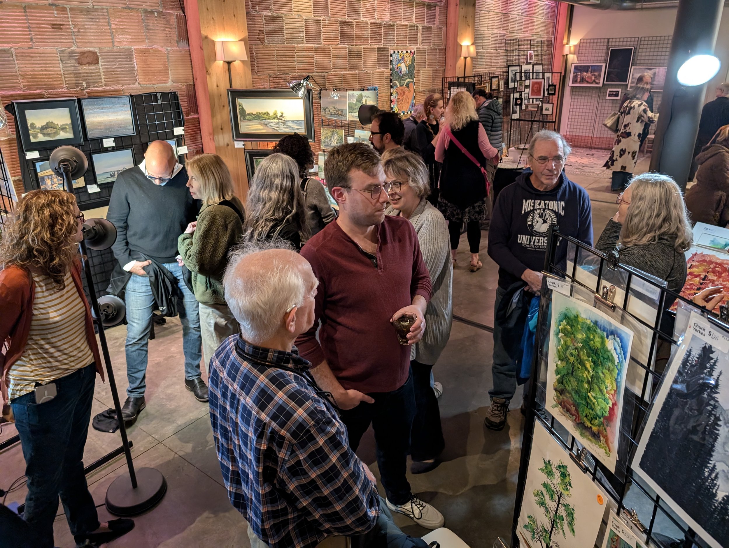 People attending an art gallery exhibition, viewing and discussing various paintings and artwork displayed on the walls and on tables.
