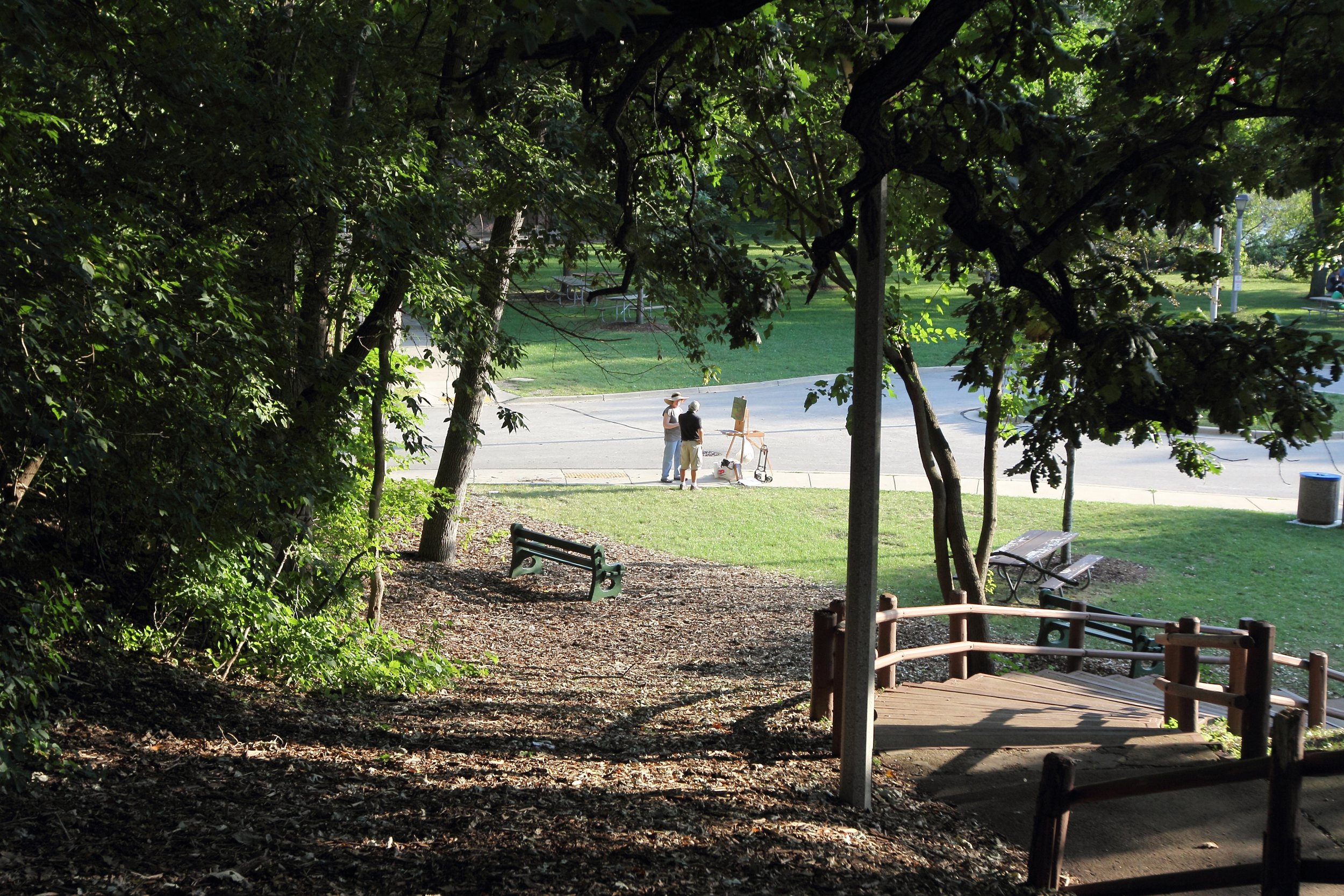 View from a shaded park trail looking toward a grassy area where two people are standing near an artist's easel on a sidewalk, with benches and trees in the background.