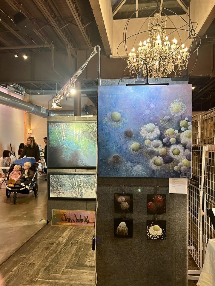 Art exhibit featuring abstract and nature-inspired paintings, with a chandelier overhead and people in the background at an indoor event.