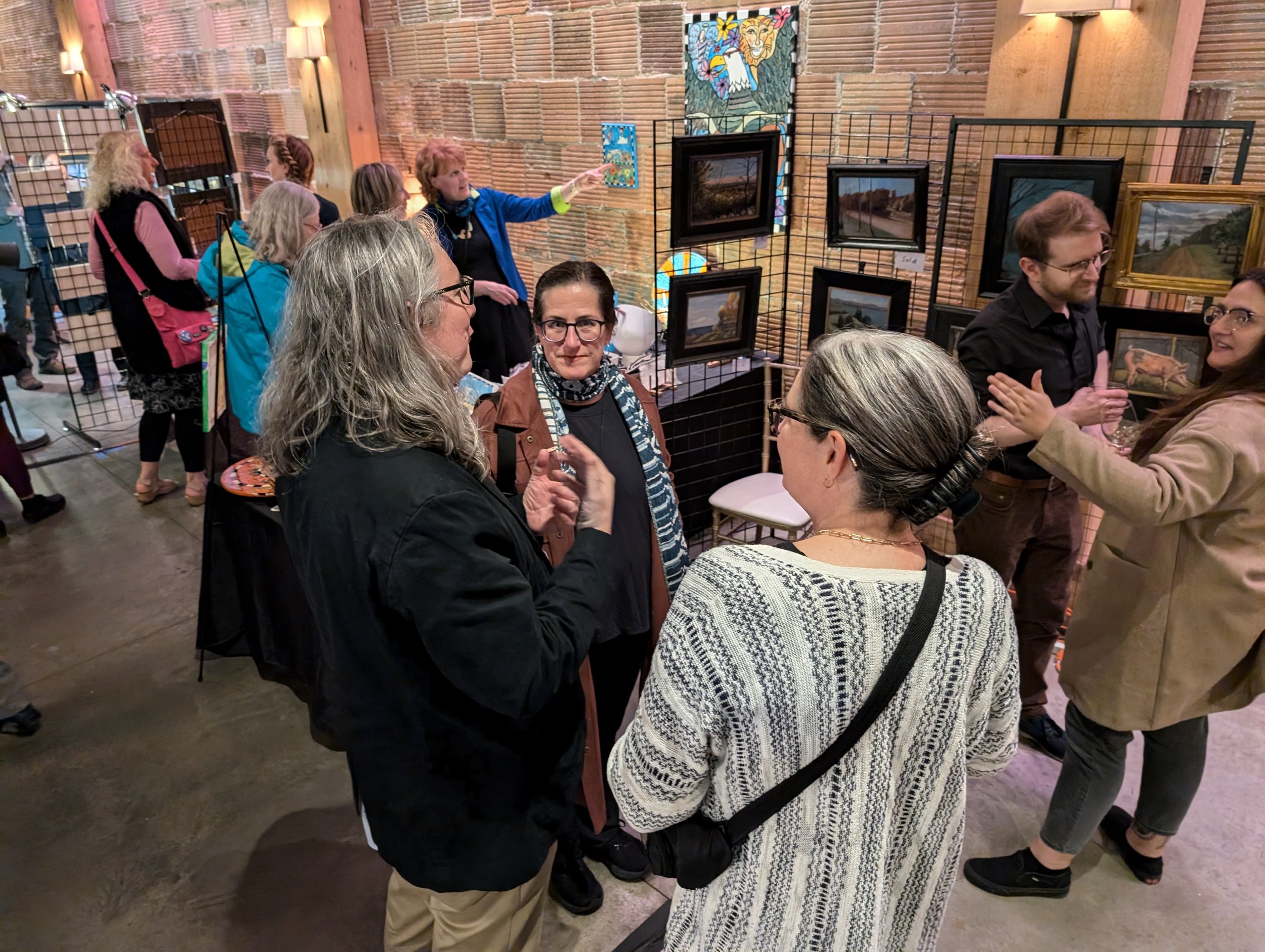 People socializing at an art gallery with framed landscape paintings and one mosaic artwork on brick walls