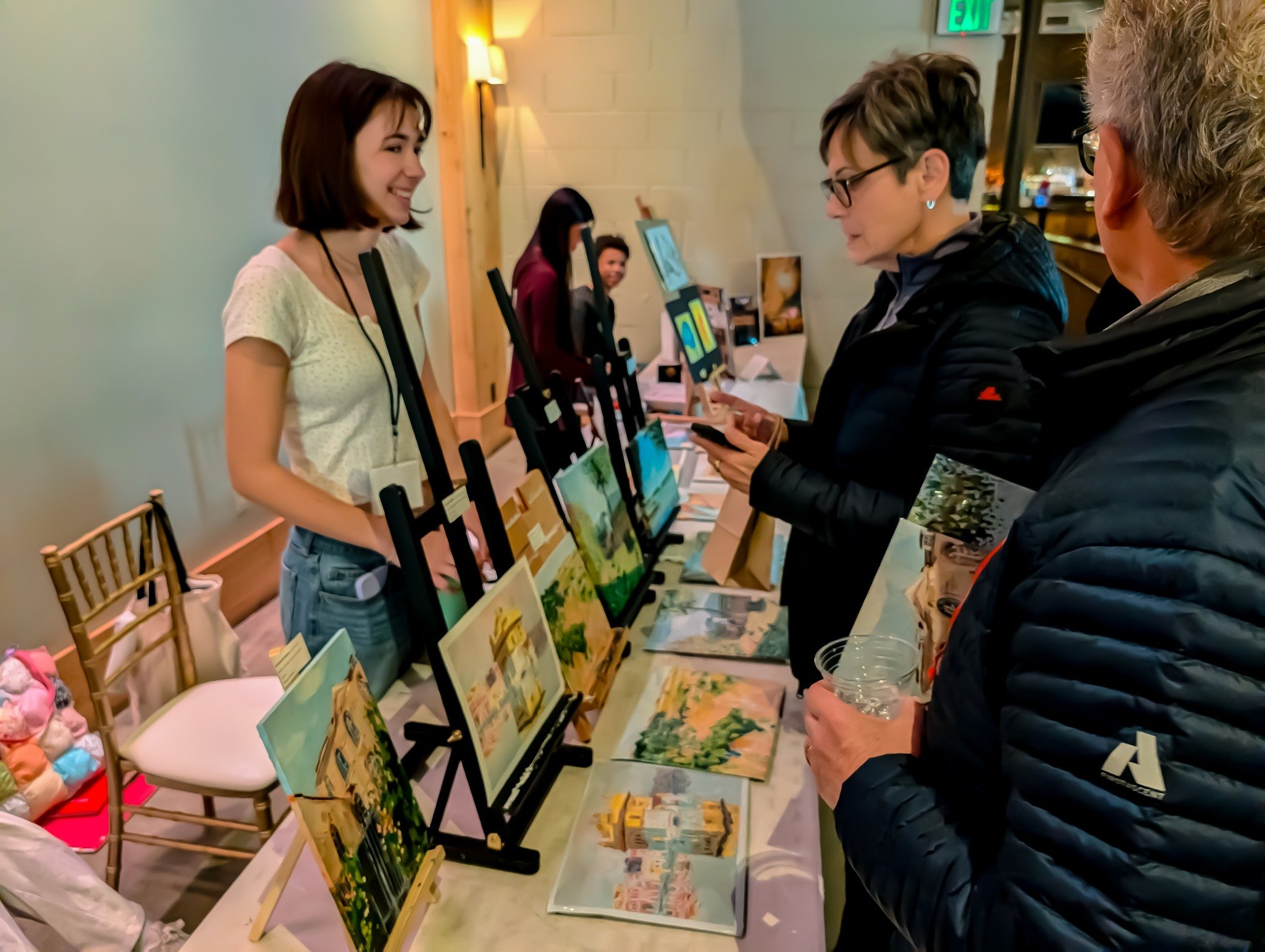A woman is smiling and talking to two customers at an art display table with paintings and greeting cards, indoors with soft lighting.