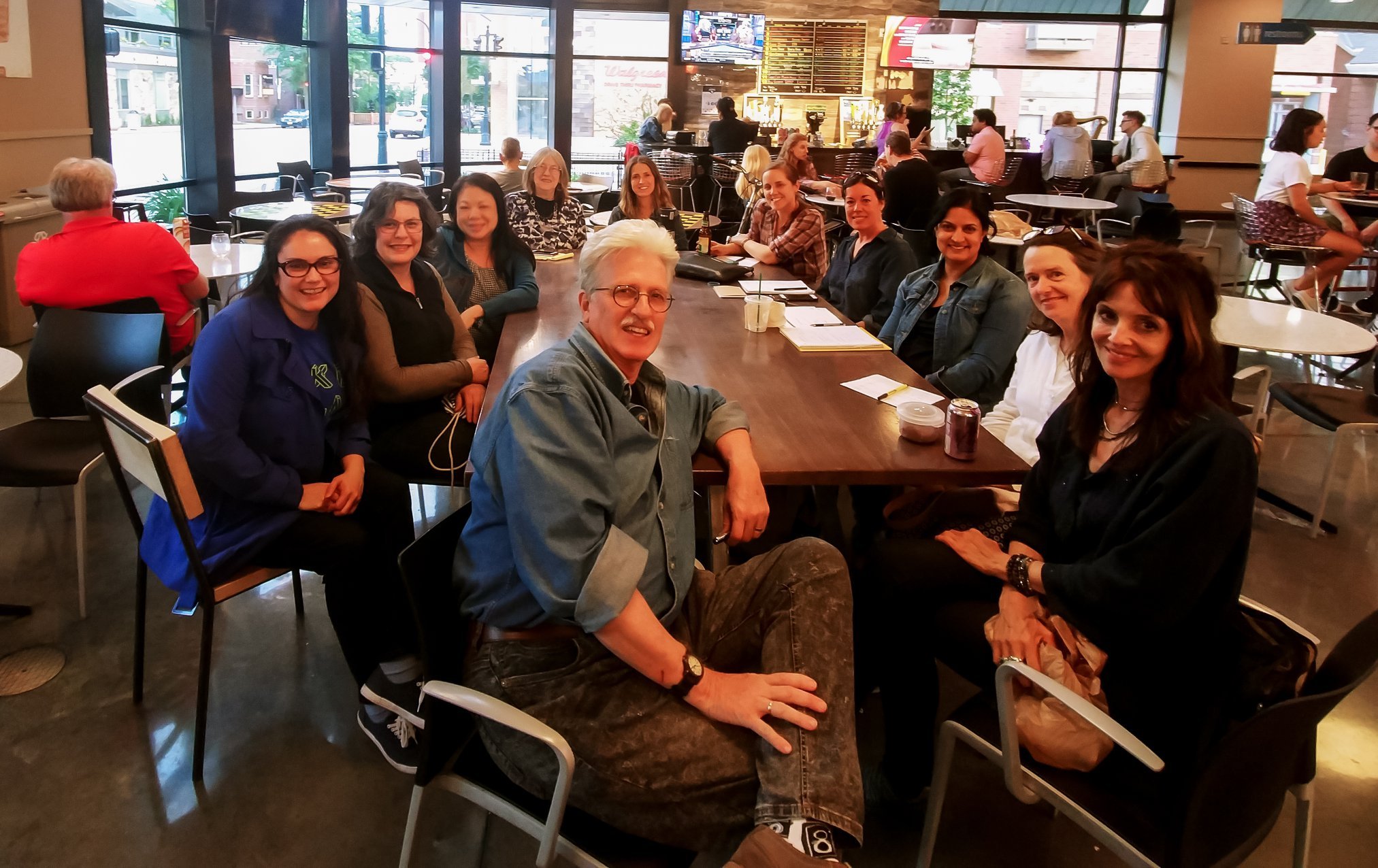 A group of people sitting around a long table in a coffee shop or cafe, smiling at the camera.