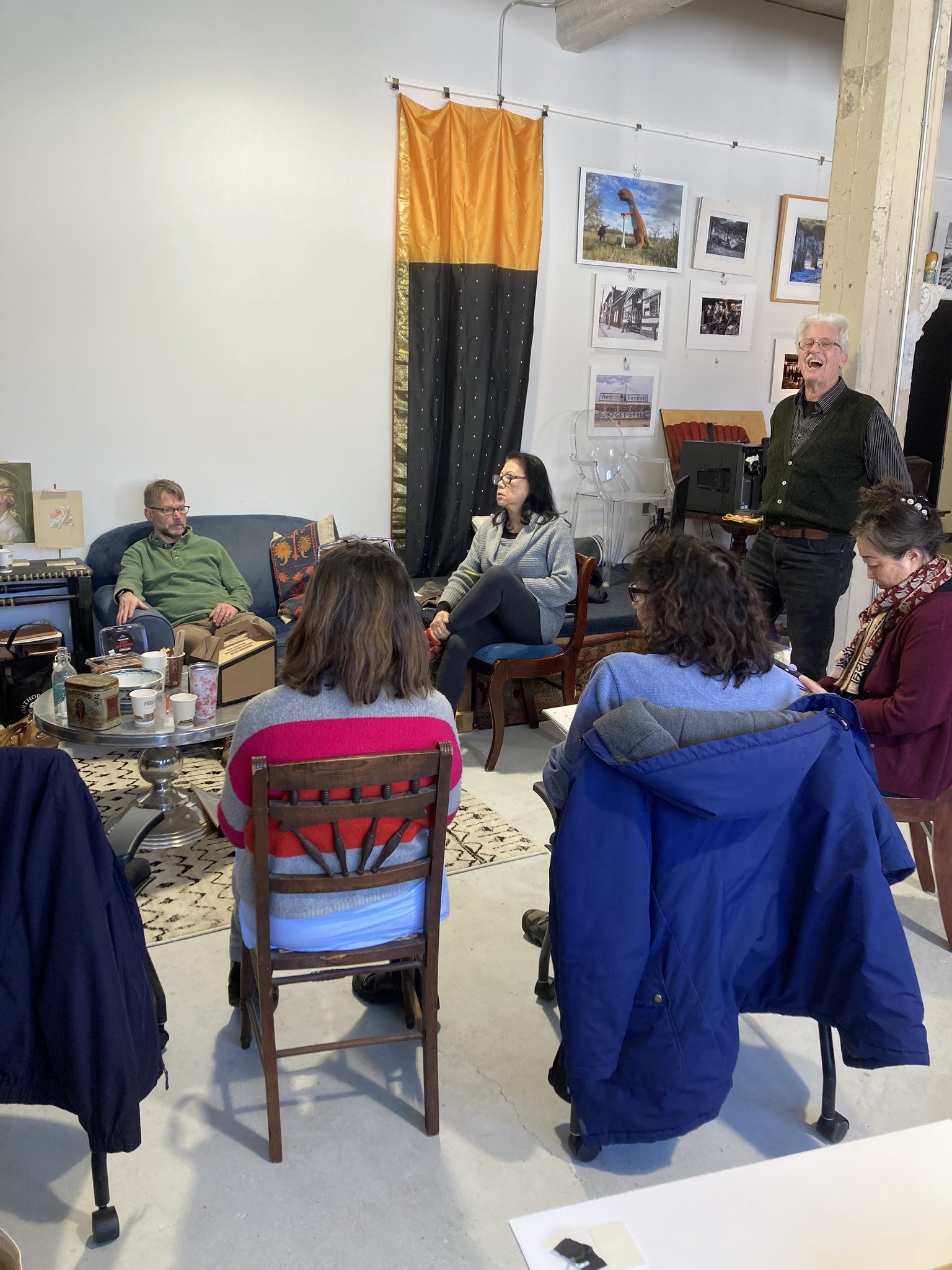 A group of people sitting and standing in a room, with one person standing near a keyboard and laughing, while others listen attentively, with framed photos and artwork on the wall behind them.