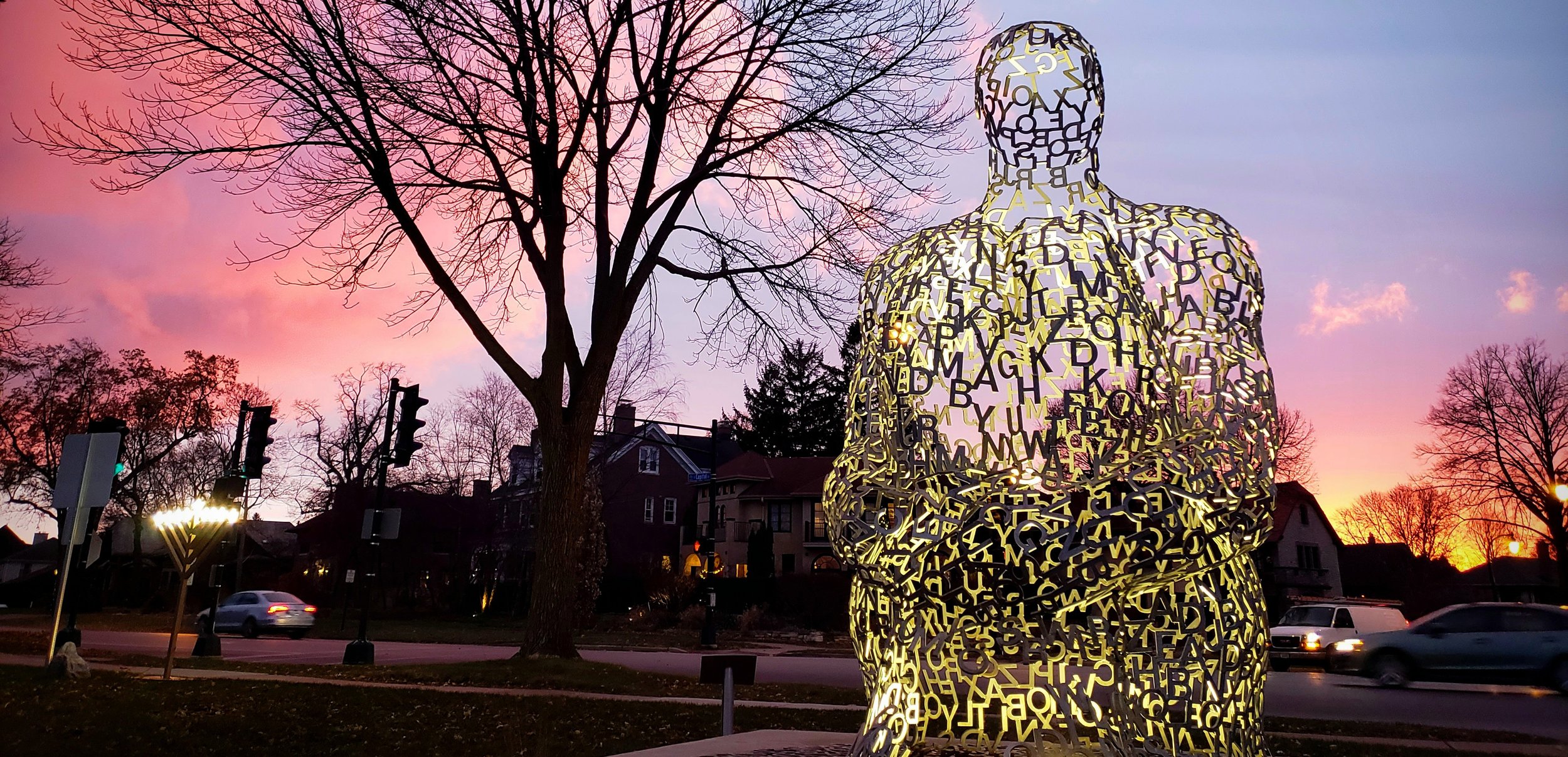A large illuminated sculpture of a human figure composed of overlapping yellow and black alphabet letters, situated outdoors at sunset with leafless trees and a neighborhood in the background.