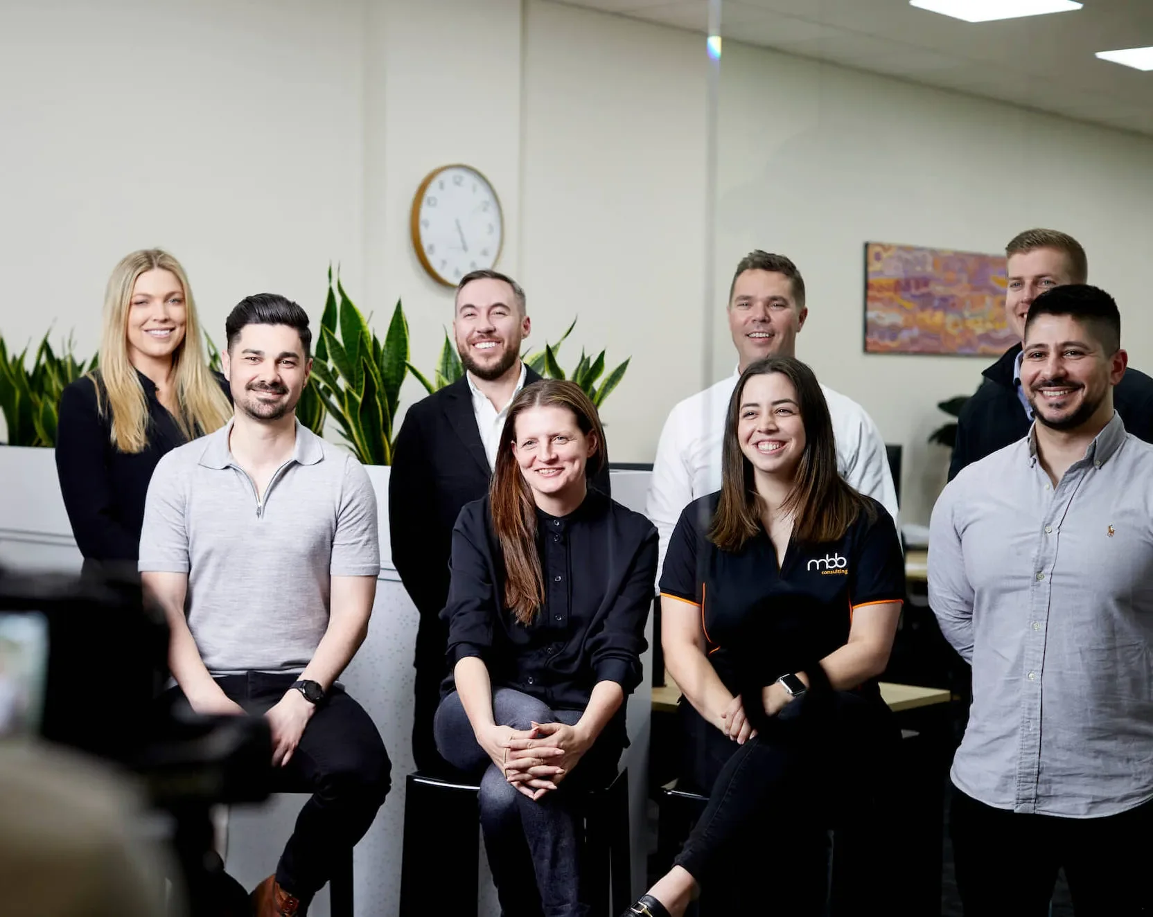 Group of diverse smiling professionals in an office setting, standing and seated in front of plants and a clock.