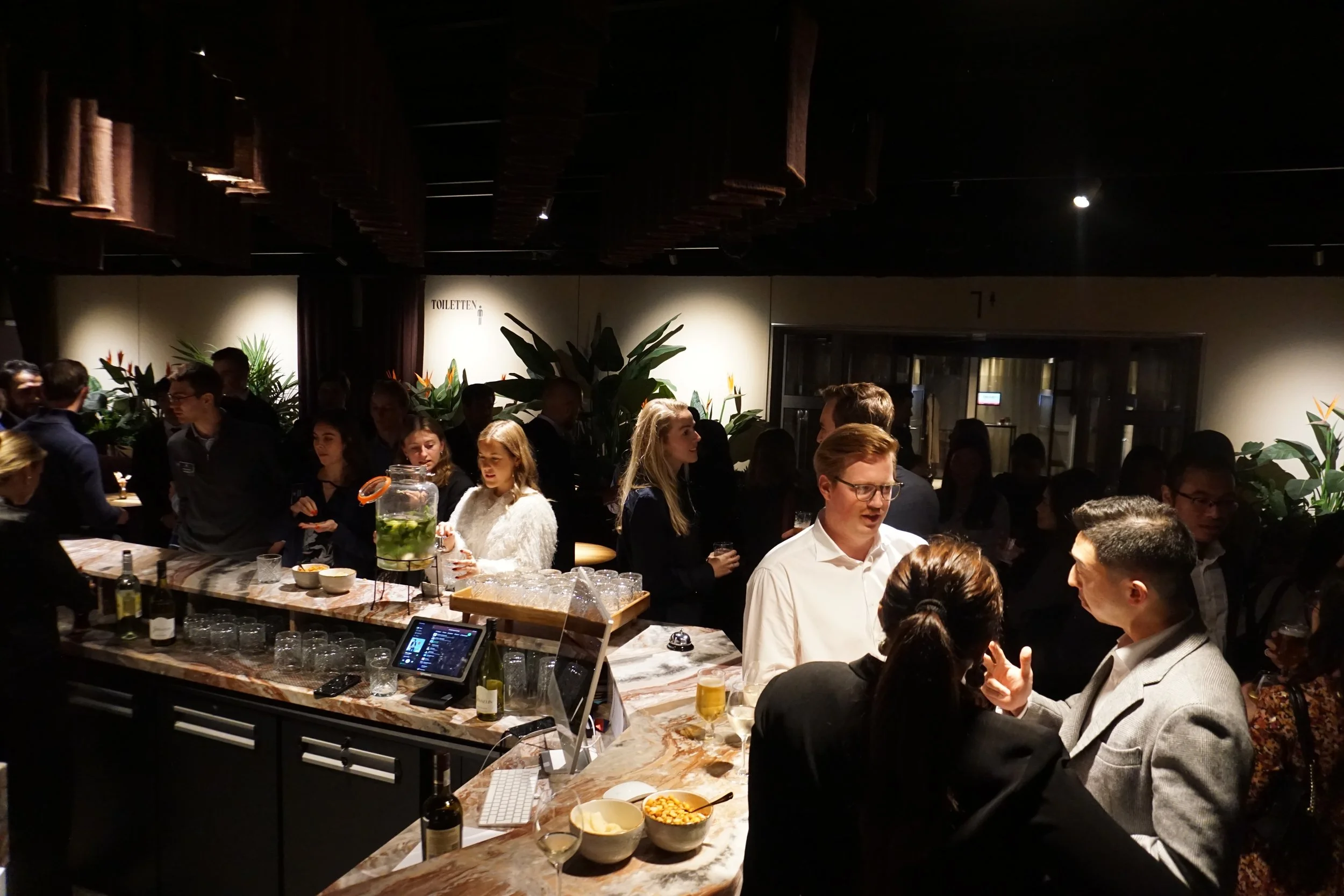 A busy indoor bar or lounge area with people socializing, some standing and some sitting, with a marble countertop, drinks, and snacks, and lush green plants in the background.