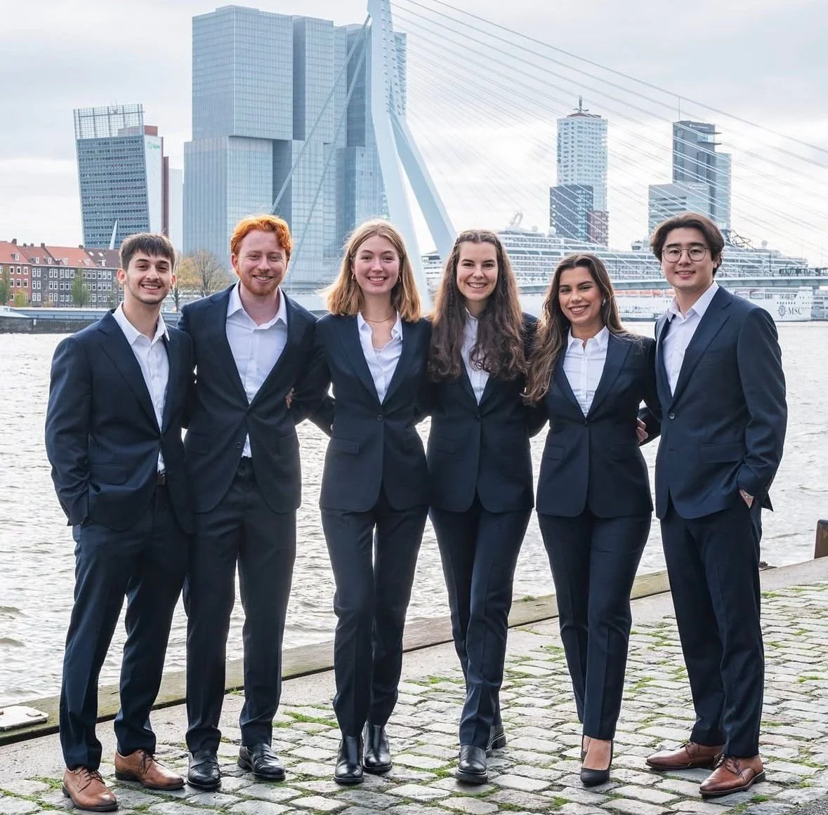 Six young adults in business suits standing together on a riverside in an urban area with modern high-rise buildings and a cruise ship in the background.