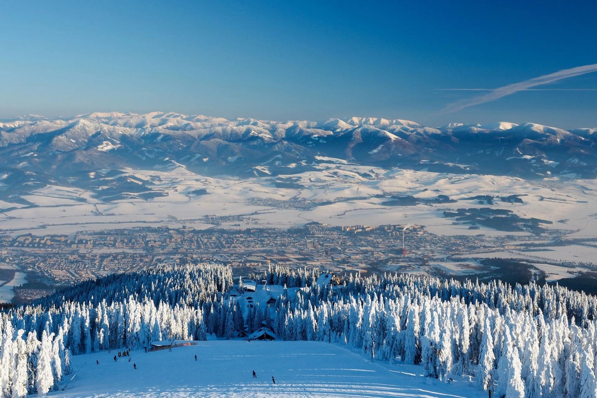 Snow-covered ski slope with skiers, surrounded by snow-laden trees, overlooking a valley with buildings, fields, and distant snow-capped mountains under a clear blue sky.