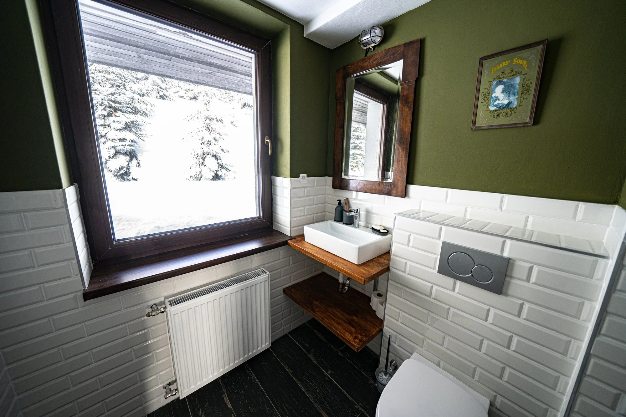 Small bathroom with a large window showing snow-covered trees outside, a white rectangular sink on a wooden countertop, a mirror framed in dark wood above the sink, dark wood flooring, white tile walls, a modern toilet with a wall-mounted flush panel, and a framed picture on the green wall.