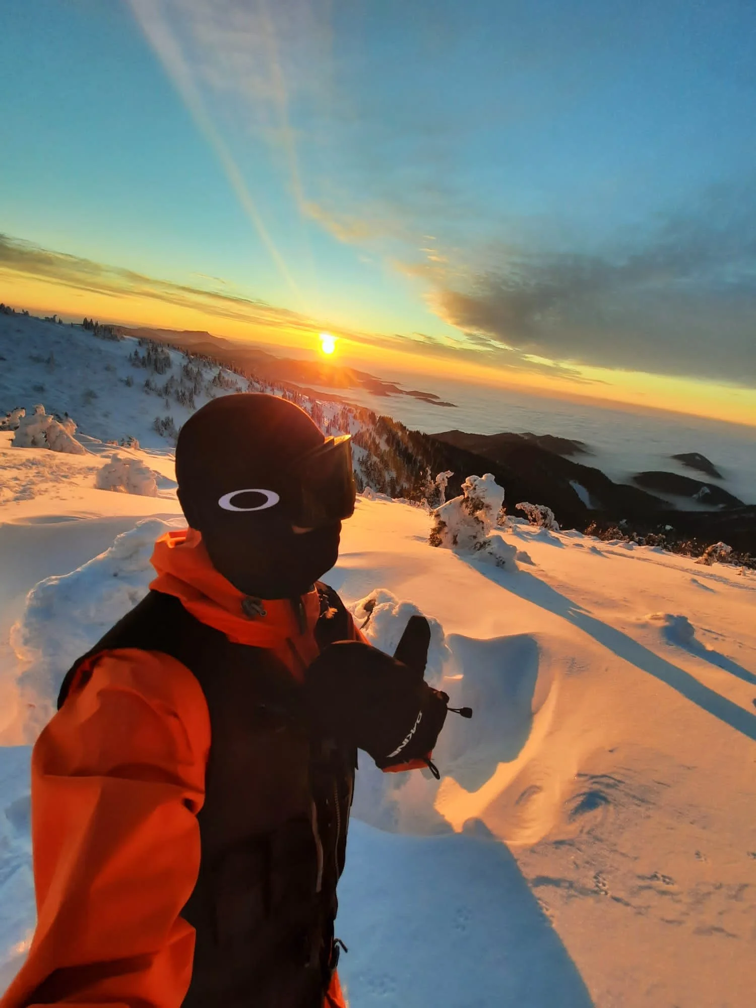 A person in winter gear, wearing a black helmet and an orange and black jacket, standing in snow with a scenic sunset or sunrise over snowy mountains and clouds in the background.