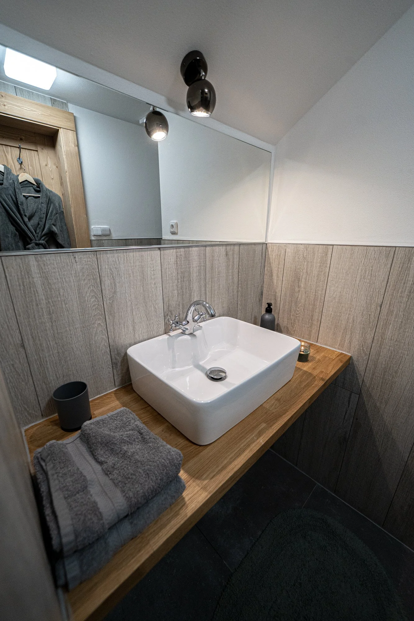Bathroom sink with a white square basin on a wooden countertop, above a dark tiled floor, with a gray soap dispenser, candle, cup, and folded towel nearby. A large wall mirror reflects part of the room and ceiling lights.
