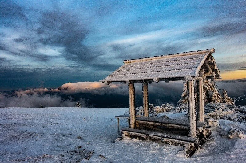 A snow-covered wooden shelter on a mountain with a dramatic cloudy sky overnight.