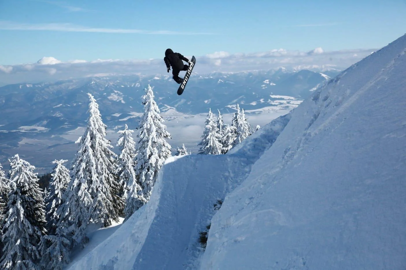 A person snowboarding off a large snow-covered ledge in a winter mountain landscape with snow-covered trees and distant mountains in the background.