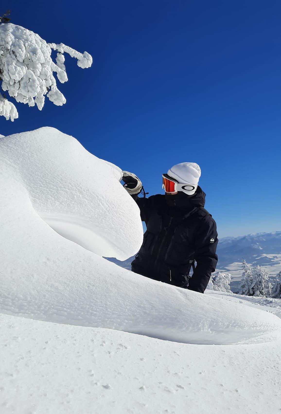 Person in black winter gear and ski goggles kneeling in deep snow in a snowy landscape with mountains in the background.
