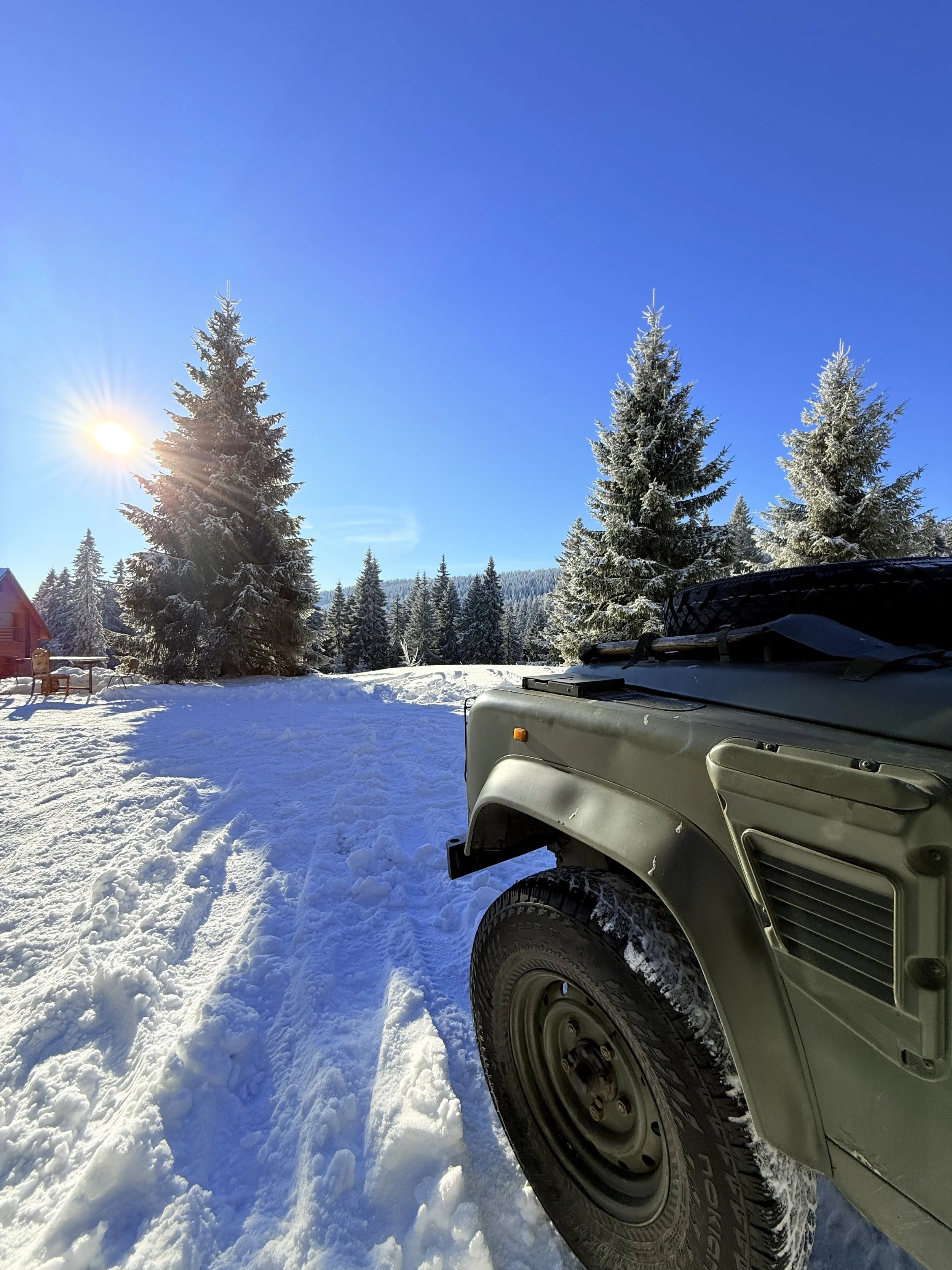 Snow-covered landscape with tall pine trees, a bright sun shining in a clear blue sky, and part of a military vehicle or jeep on the right in the foreground.