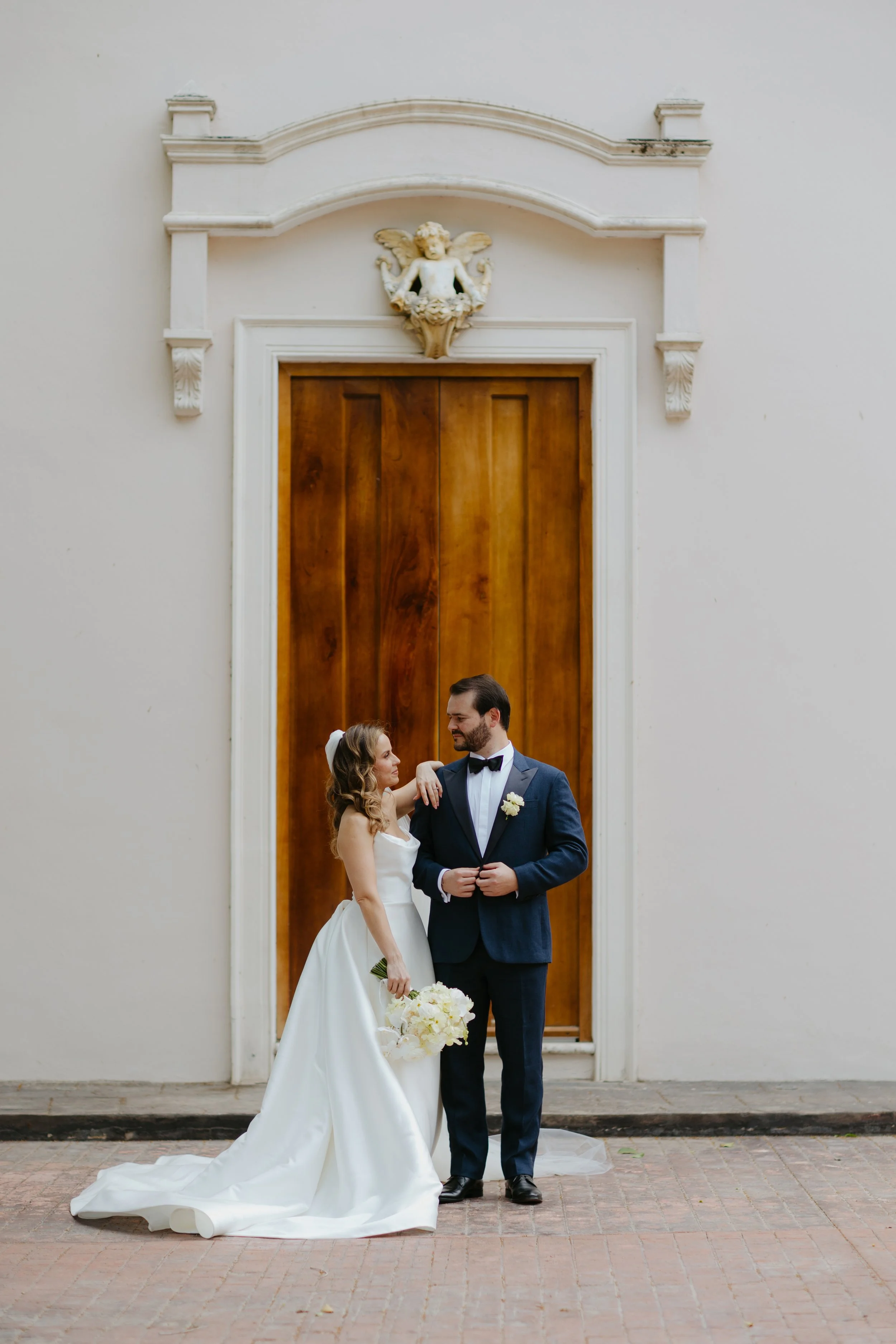 Bride and groom standing in front of a large wooden door with ornate white molding and angel sculpture above, during wedding day.