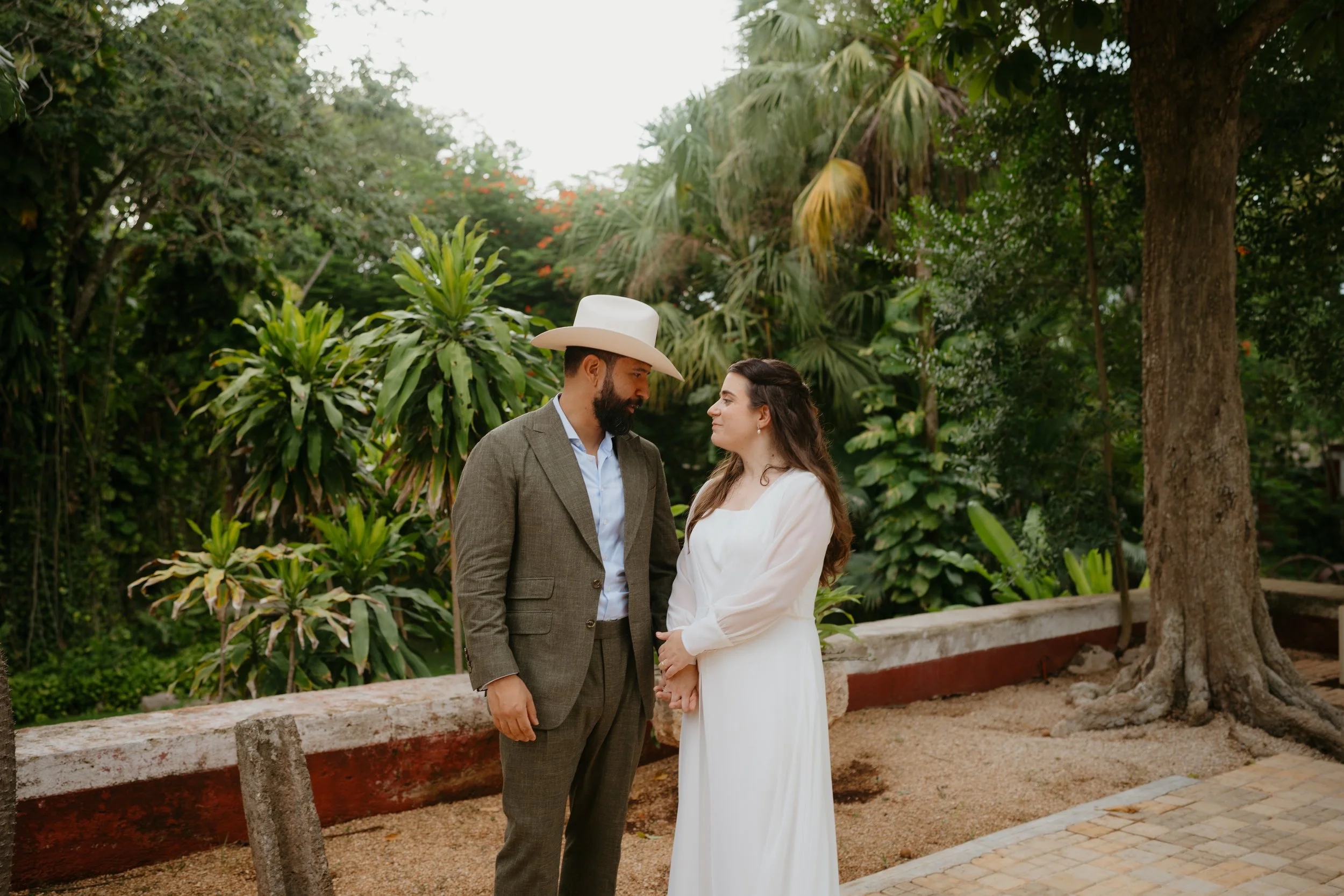 A couple dressed in formal attire standing close together outdoors, holding hands, surrounded by lush green trees and plants.