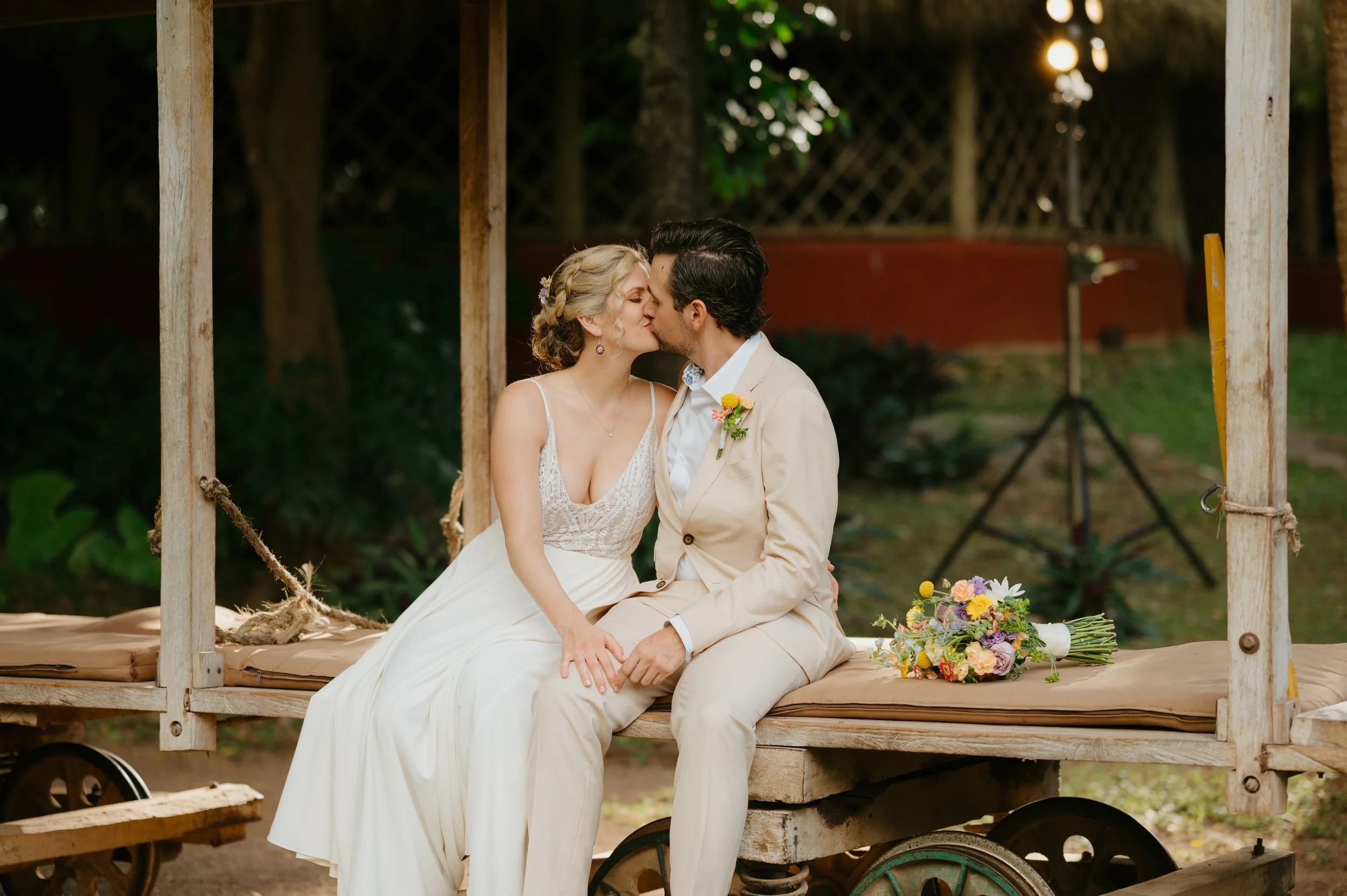 A bride and groom sitting on a wooden cart, kissing, outdoors during their wedding, with a bouquet of flowers beside them.