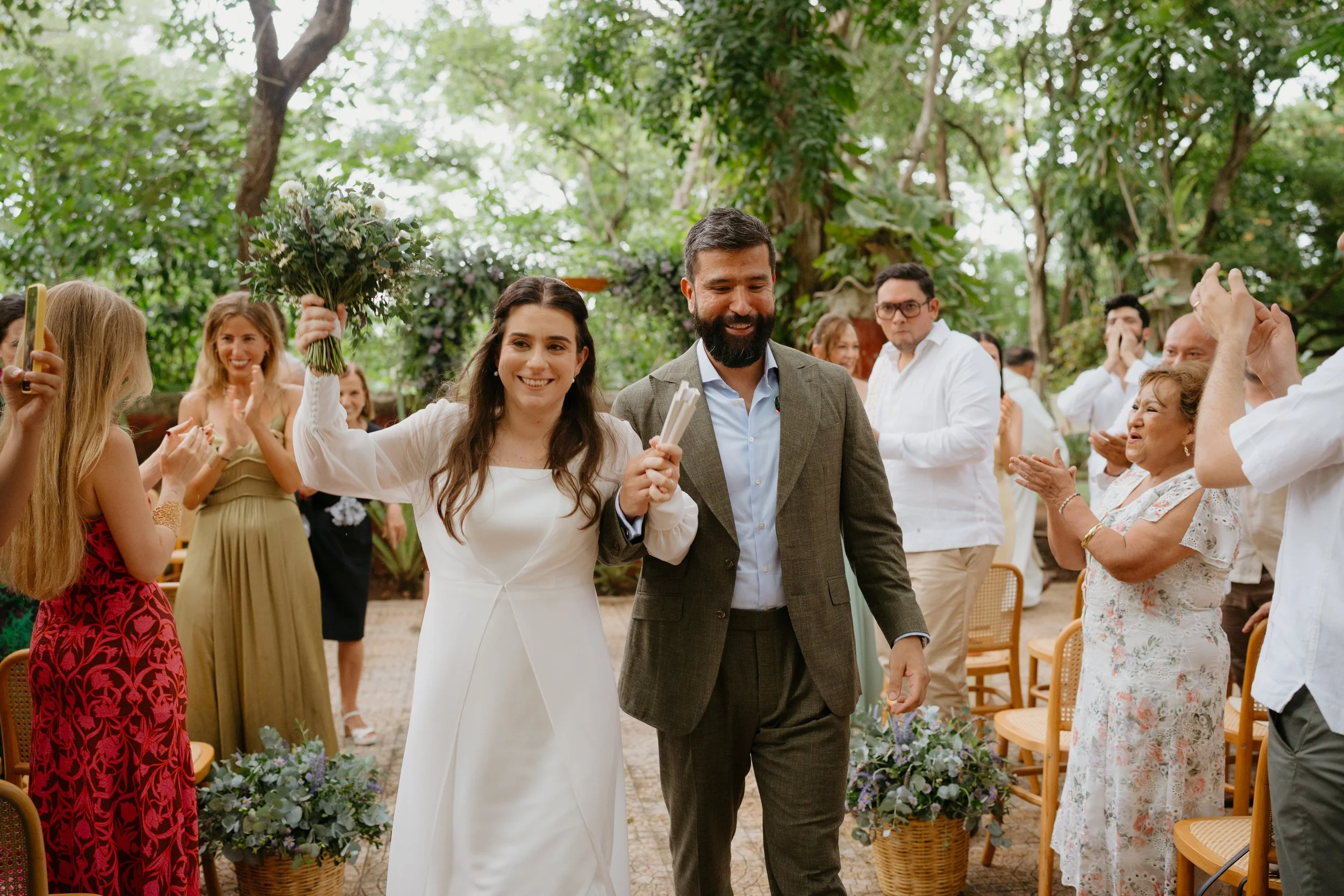 Bride and groom walking down the aisle at their wedding, smiling and holding hands, surrounded by smiling guests in an outdoor garden setting.