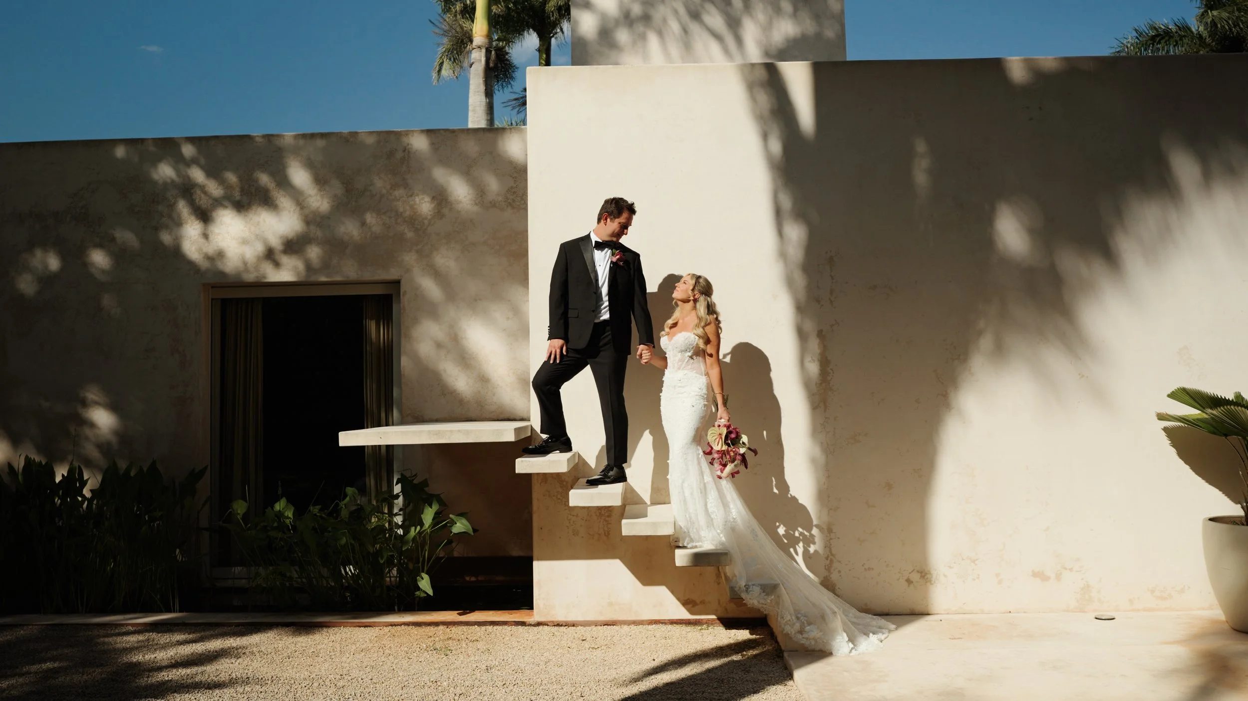 A couple dressed in wedding attire standing outdoors, smiling and embracing with a lush green background.