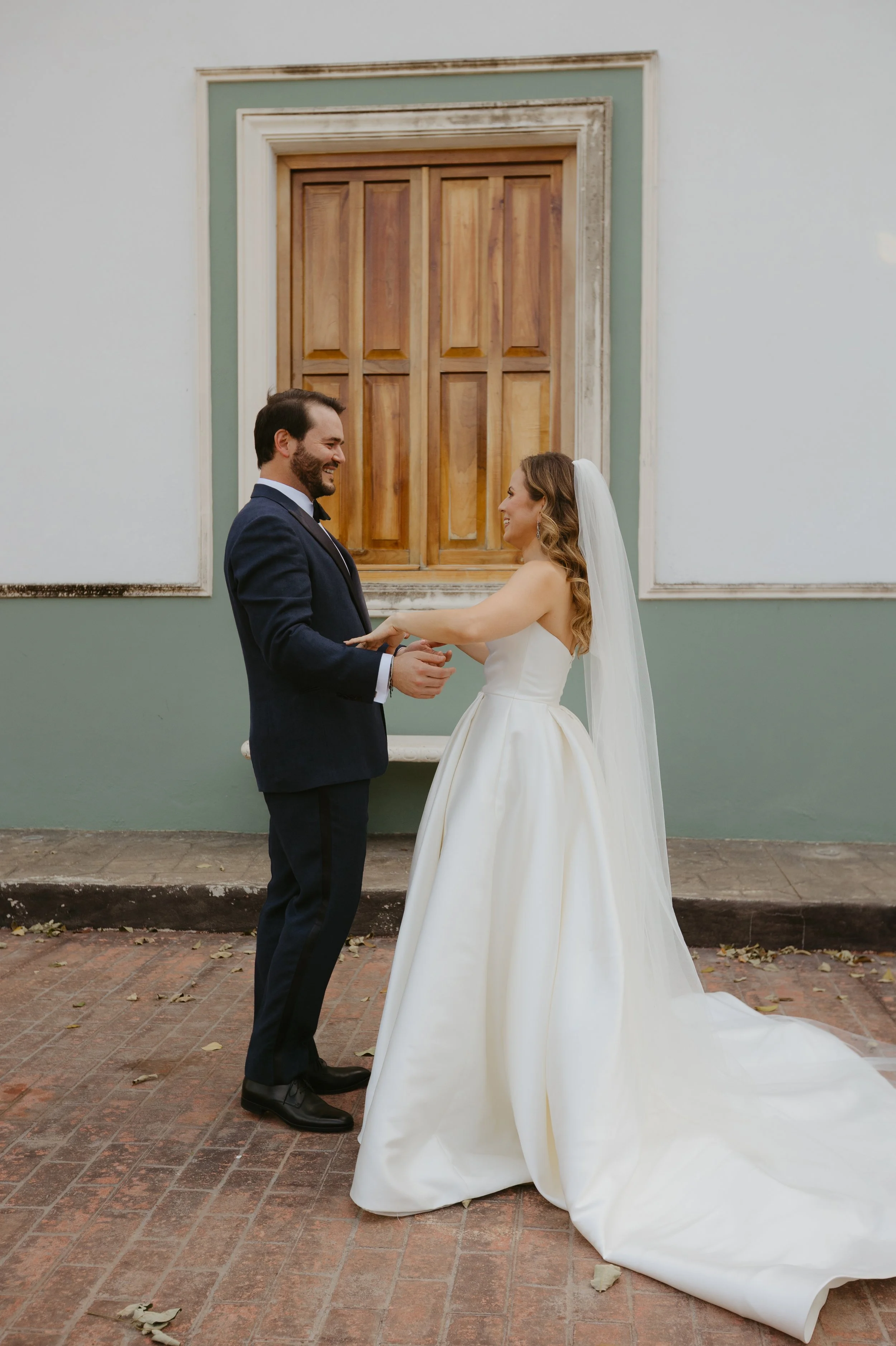 A bride and groom holding hands and smiling at each other outside, with a green and white wall and wooden window behind them.