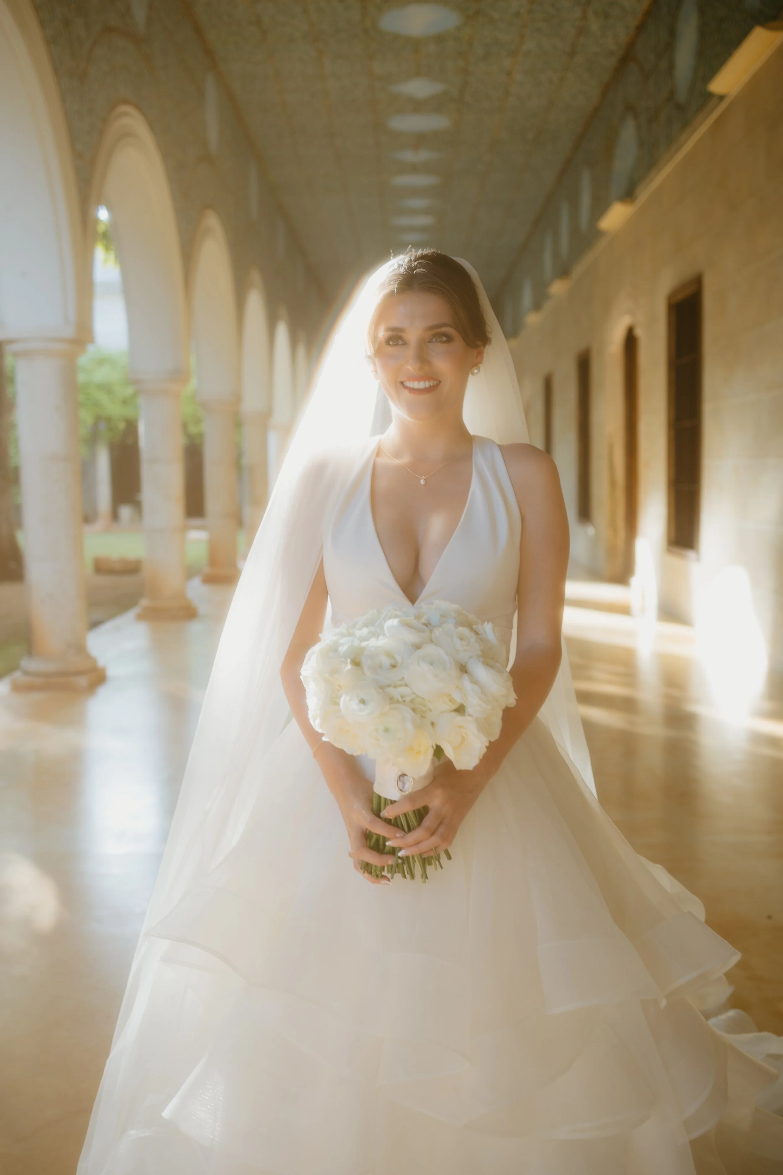 Bride in white wedding dress holding a bouquet of white flowers, standing under an arched corridor with sunlight streaming from behind.