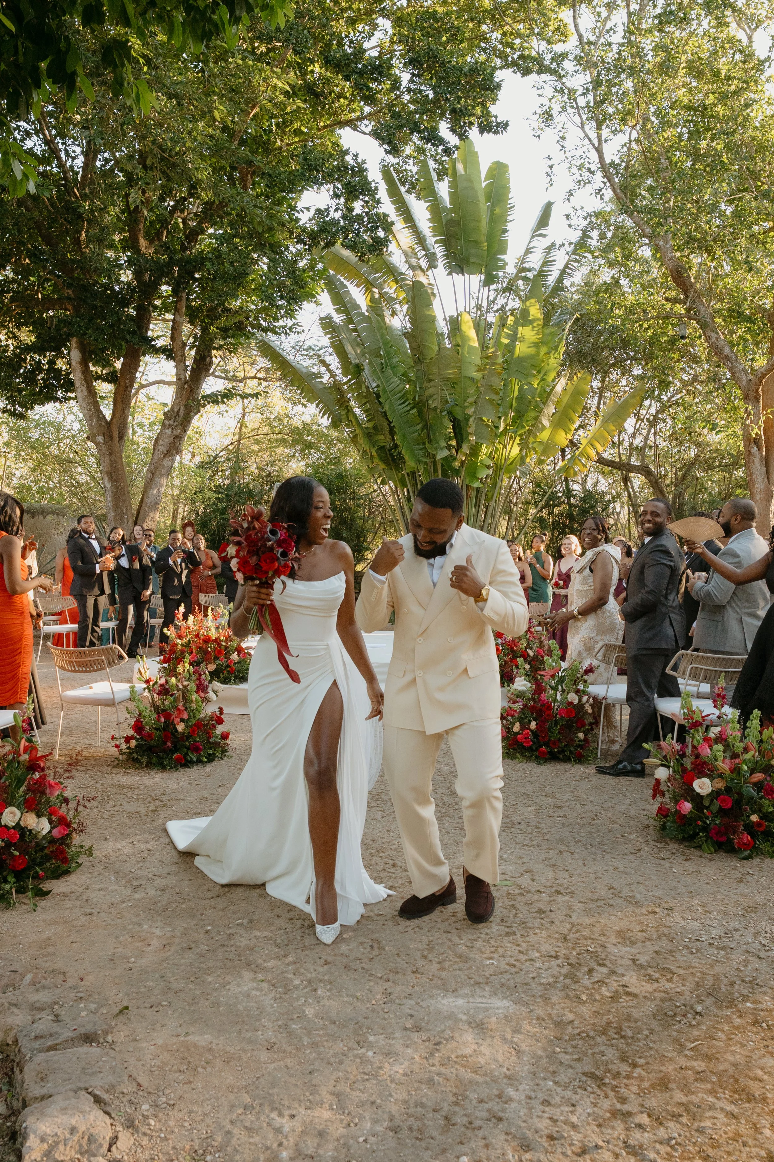 A bride in a white wedding dress and a groom in a light-colored suit celebrating during a wedding ceremony outdoors, surrounded by guests and lush greenery.