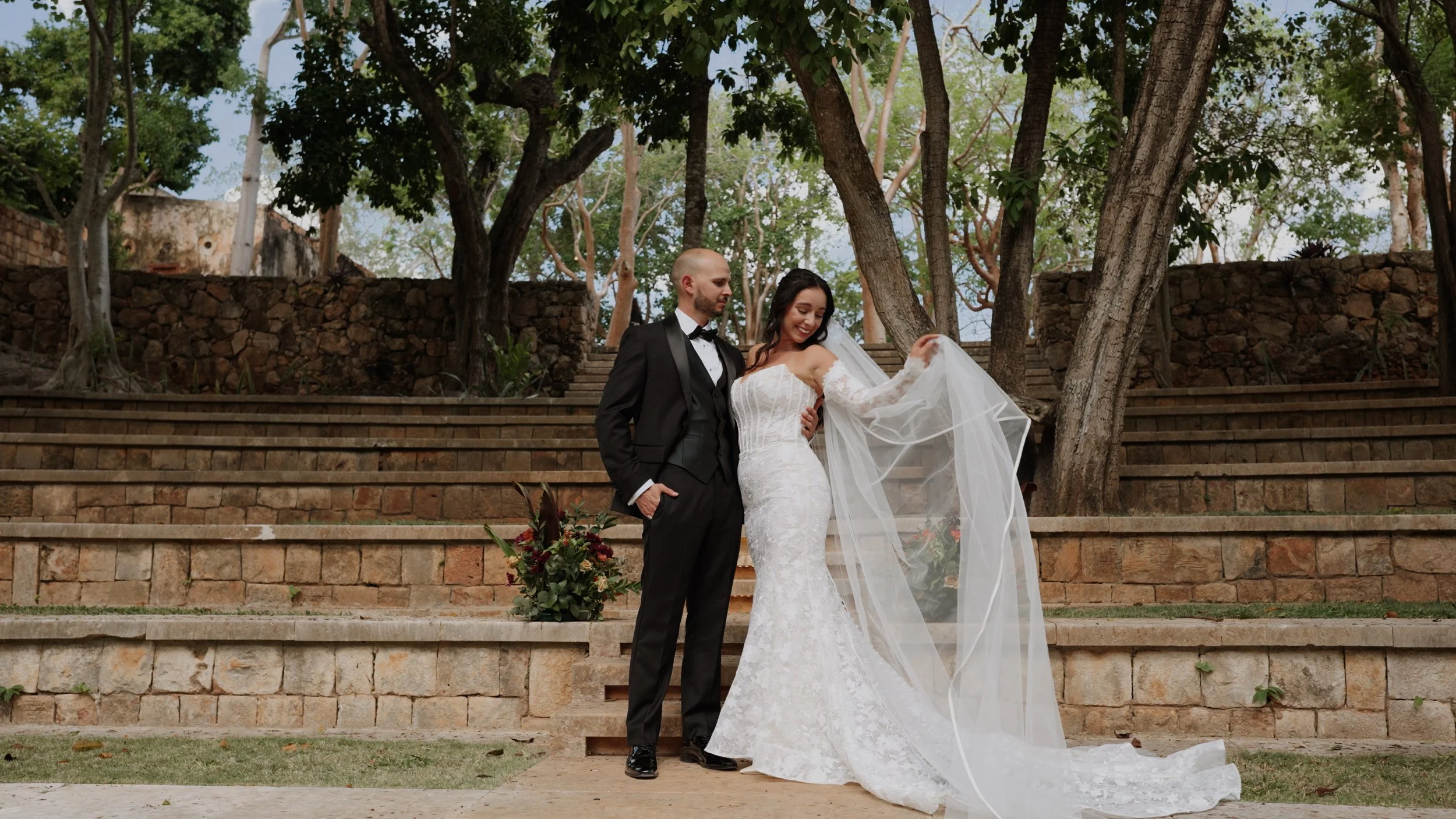 A bride and groom share a kiss outdoors during their wedding ceremony, with guests in the background and a large house and trees in the distance.