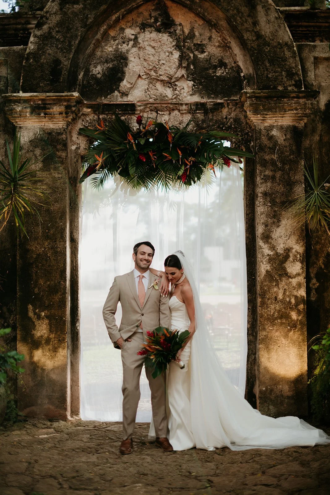 A bride and groom share a kiss outdoors during their wedding ceremony, with guests in the background and a large house and trees in the distance.