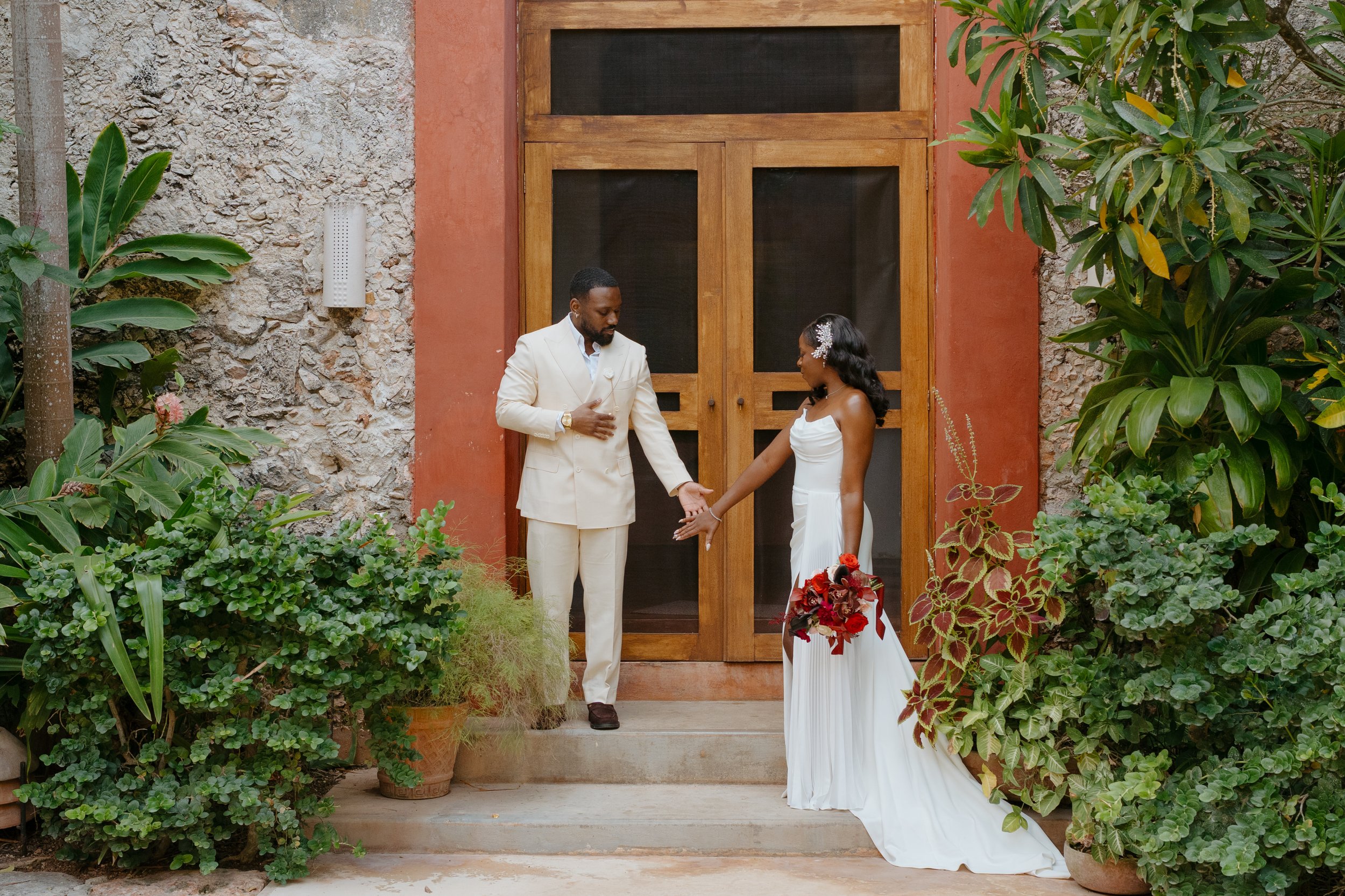 A man in a white suit and a woman in a white gown holding a bouquet of red flowers stand on steps outside a rustic building with wooden doors, surrounded by green plants.