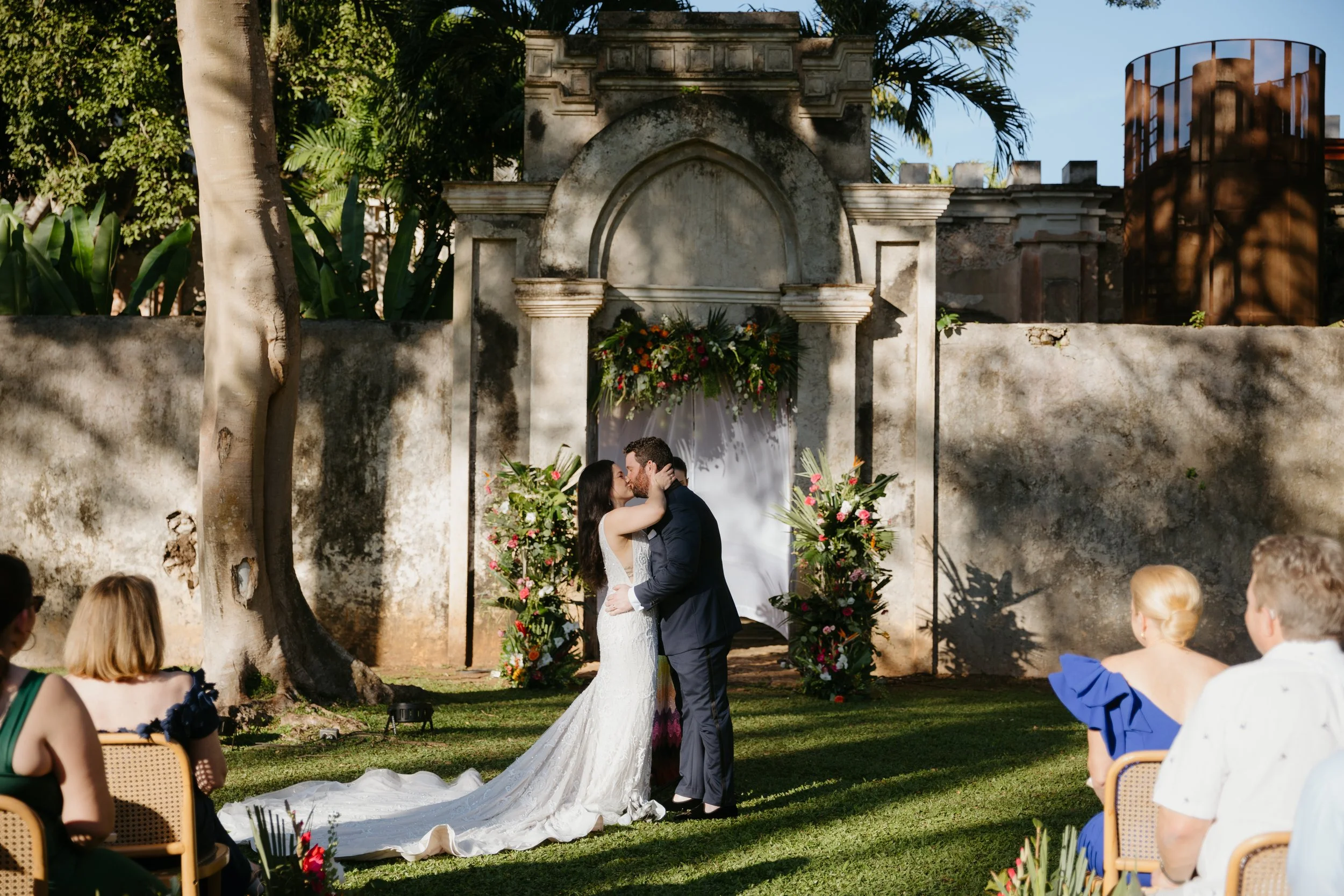 A wedding ceremony taking place outdoors at a historic stone wall with floral decorations, featuring a bride and groom sharing a kiss, surrounded by seated guests.