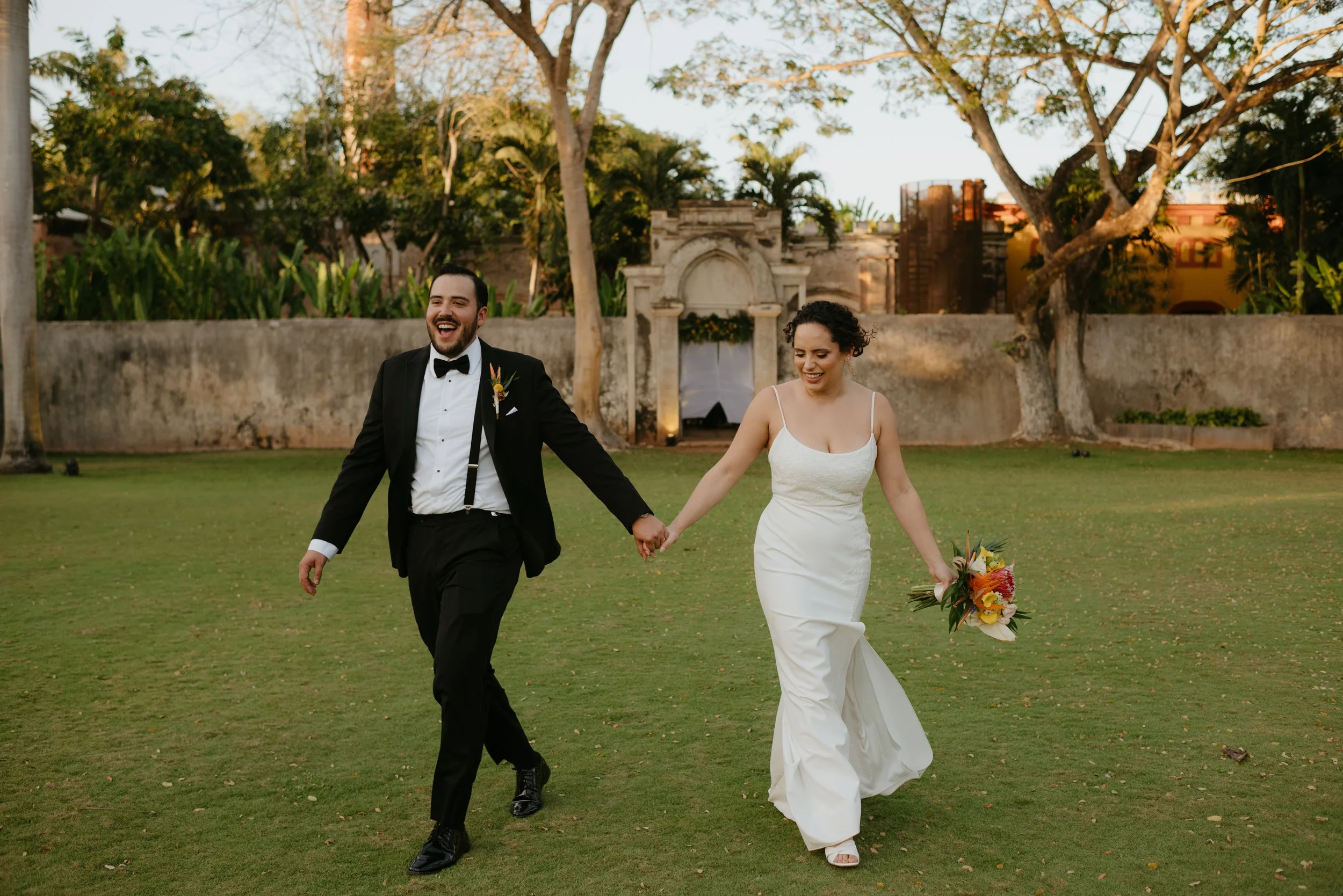 A newlywed couple holding hands and walking on a grassy lawn, smiling happily during their wedding celebration, with trees and a stone structure in the background.