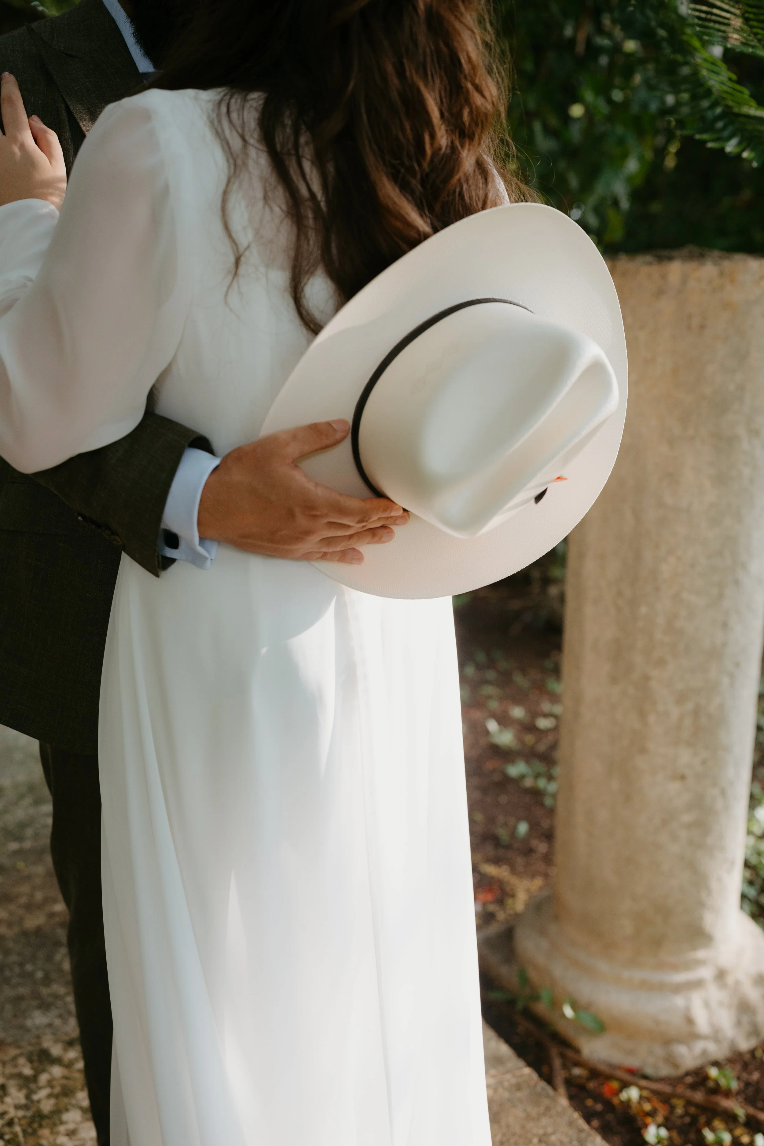 A couple embracing at a wedding, with the woman dressed in a white gown holding a white hat, and the man in a suit, next to a stone column and green foliage.