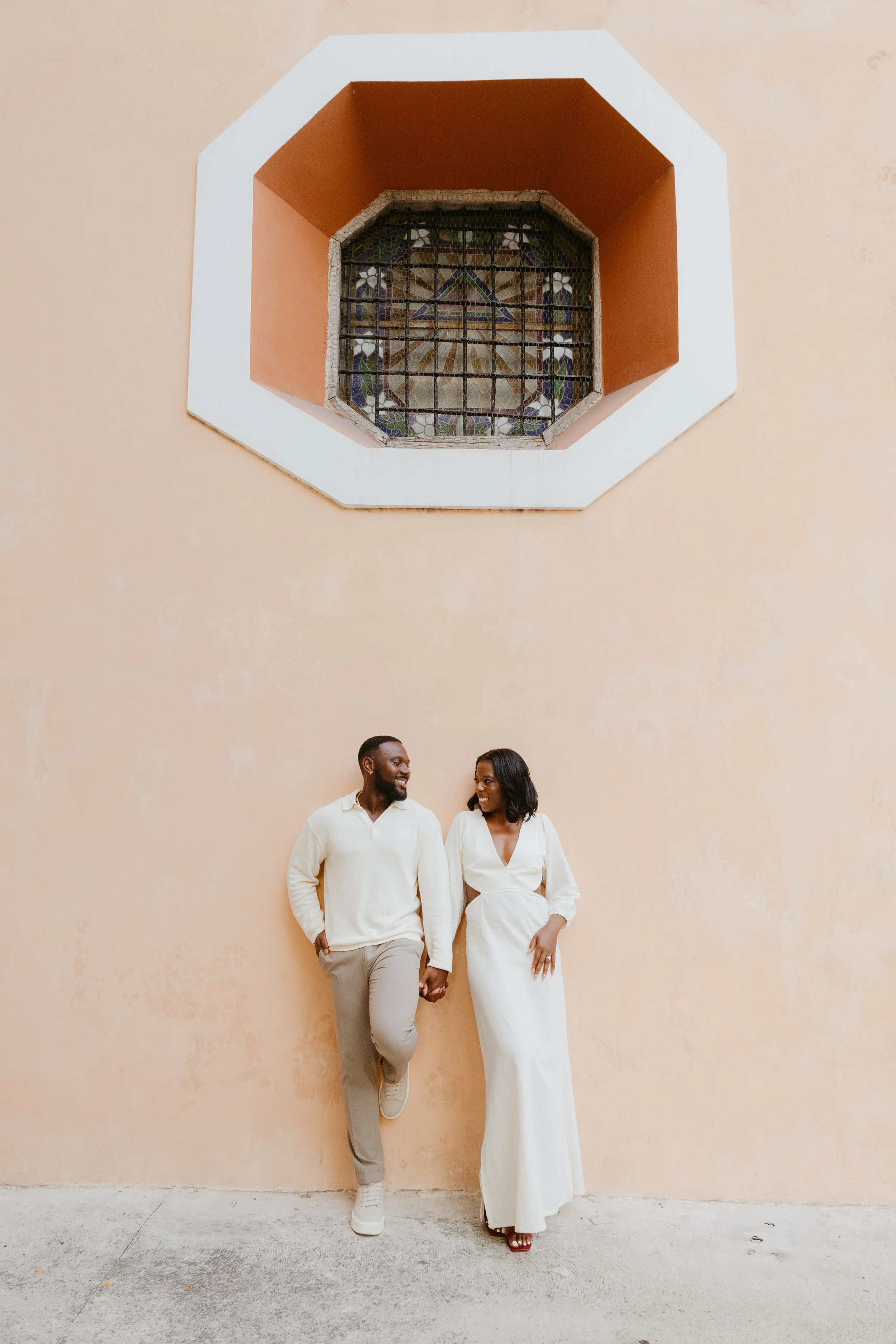 A newlywed couple standing outdoors on a grassy area near a pond, surrounded by lush green trees. The bride is holding a bouquet and wearing a white wedding dress with a long train, smiling and leaning into the groom, who is in a black suit and white shirt. They are embracing and looking at the camera.