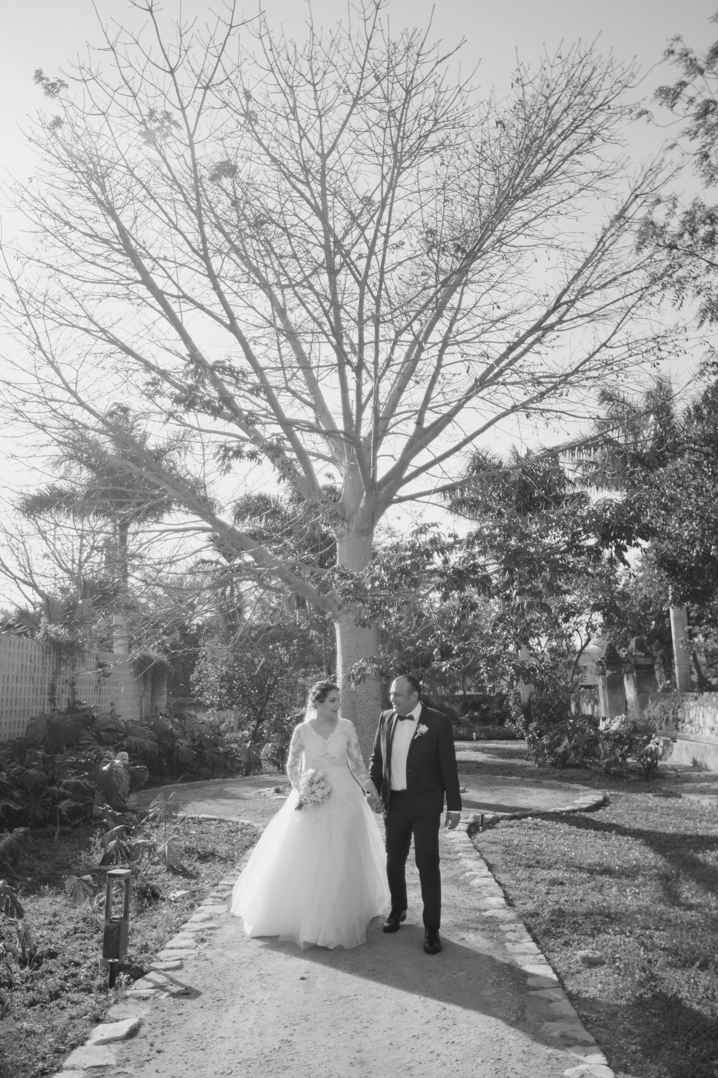 A bride and groom walking hand in hand on a garden path, surrounded by trees and greenery, during their wedding day.