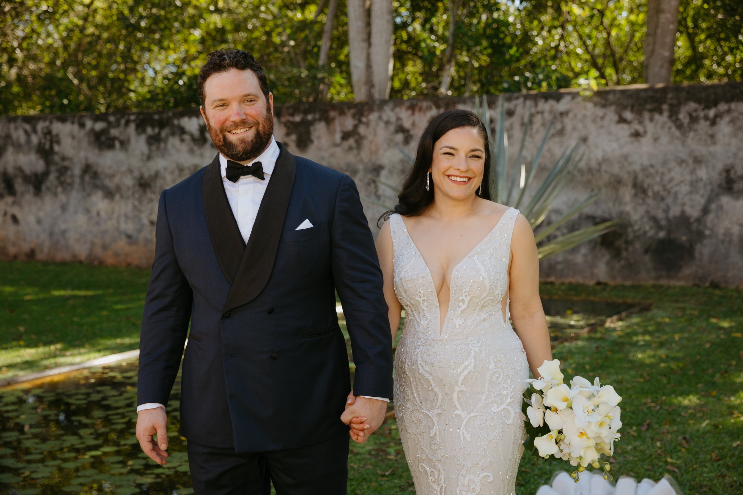 A newlywed couple holding hands outdoors, smiling, with a stone wall and greenery in the background. The man is wearing a navy tuxedo with a black bow tie, and the woman is in a white lace wedding dress holding a bouquet of white flowers.