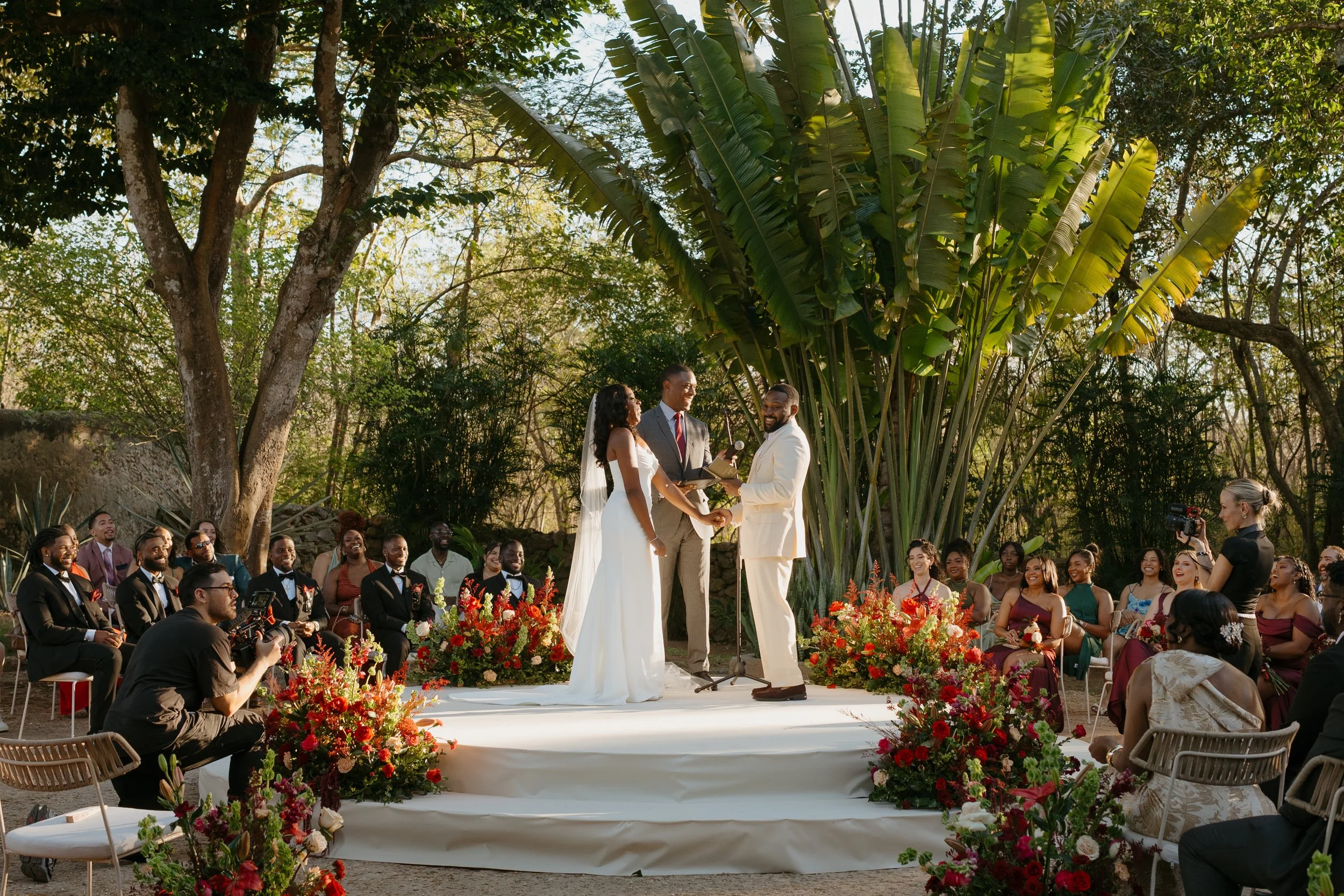 A two-tier wedding cake decorated with pink hearts, roses, and horseshoes, with a bride and groom figure topper in wedding attire, standing on the top tier.
