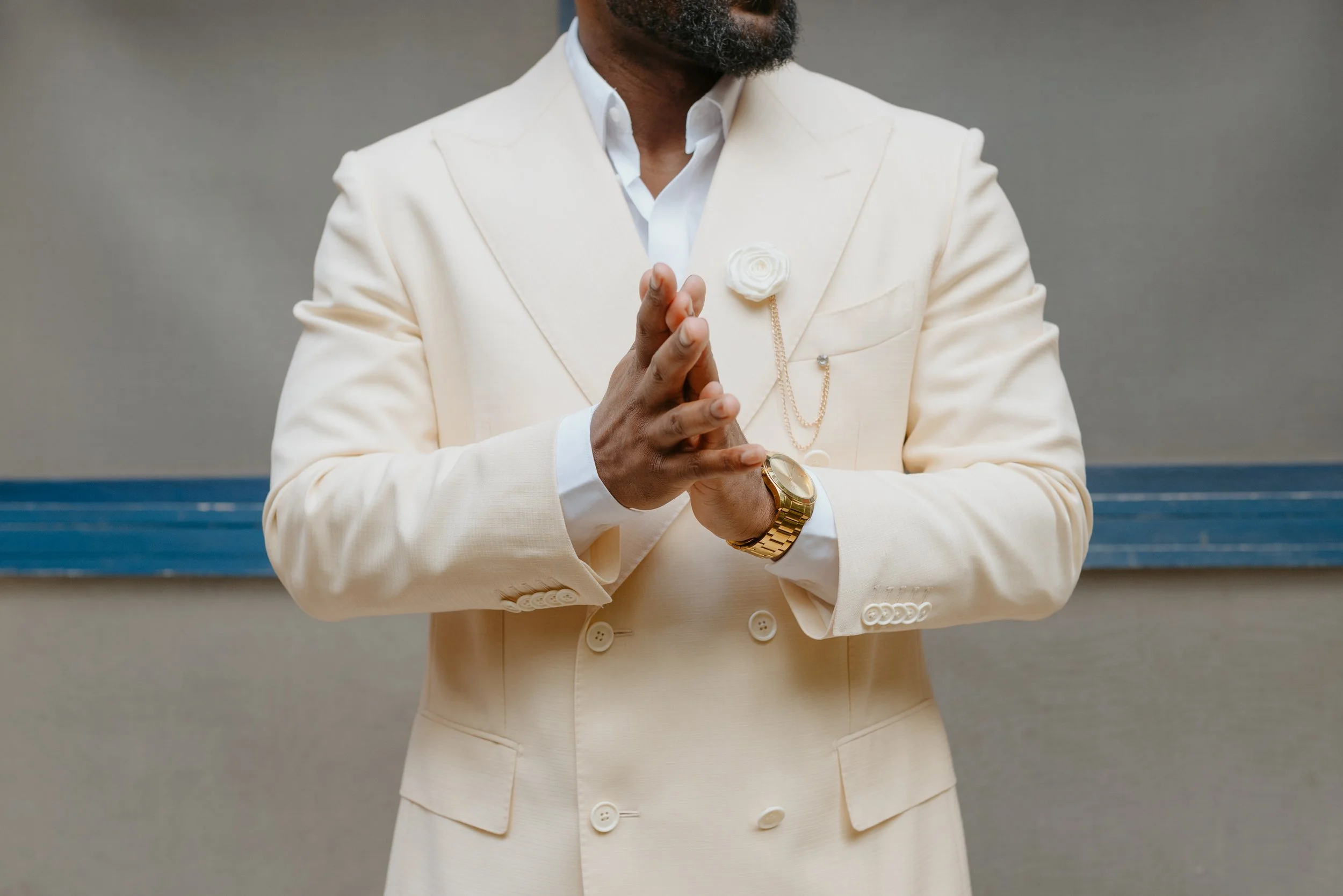 A man dressed in a cream-colored suit with a white shirt, wearing a gold watch and a white flower lapel pin. His hands are clasped together in front of his chest.