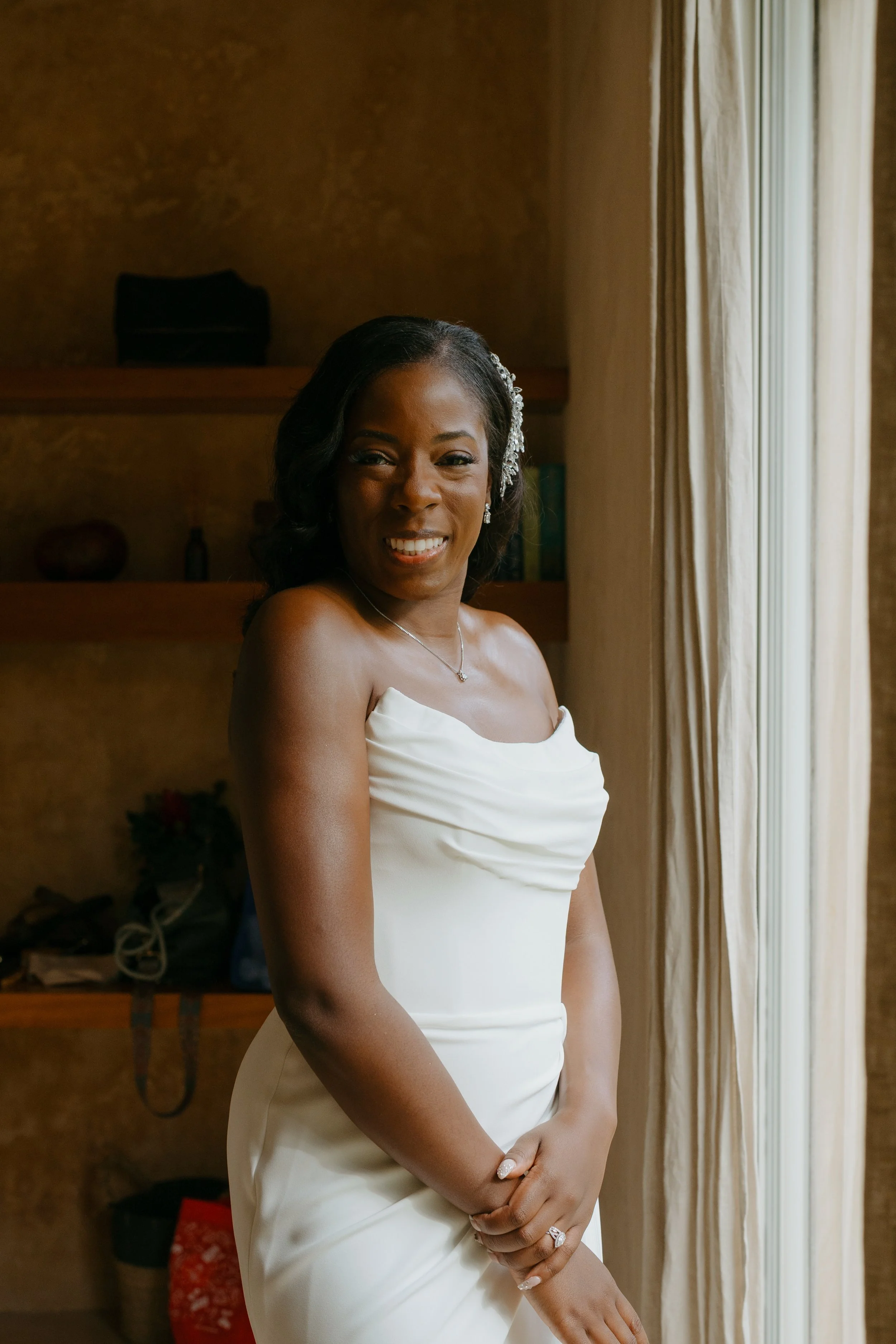 A smiling woman in a white strapless dress standing near a window with curtains, indoors.