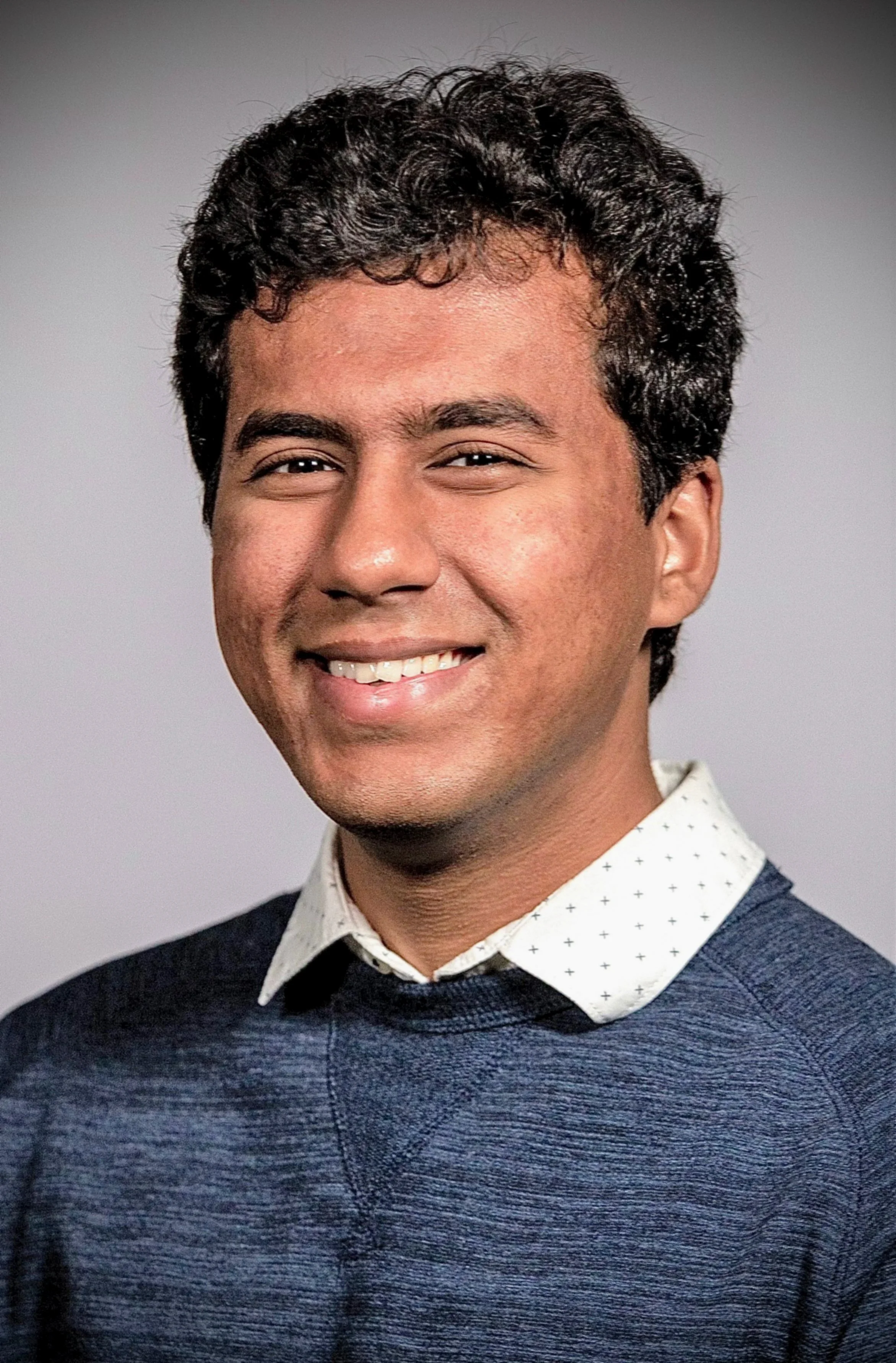 A young man with short curly black hair, smiling, wearing a dark blue sweater over a white shirt with small black dots, against a plain gray background.