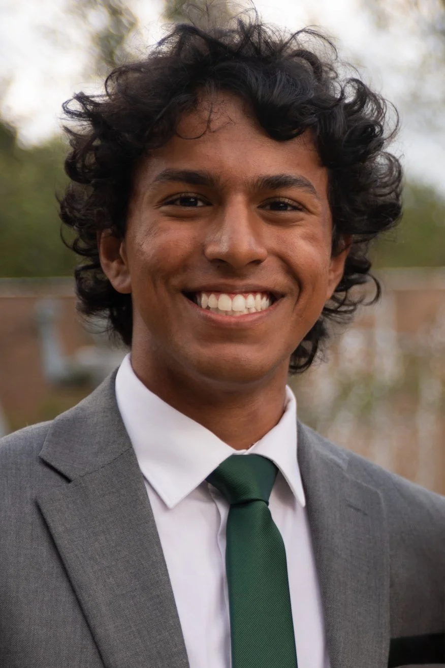 A smiling young man wearing a gray suit, white shirt, and green tie outdoors with blurred trees and buildings in the background.