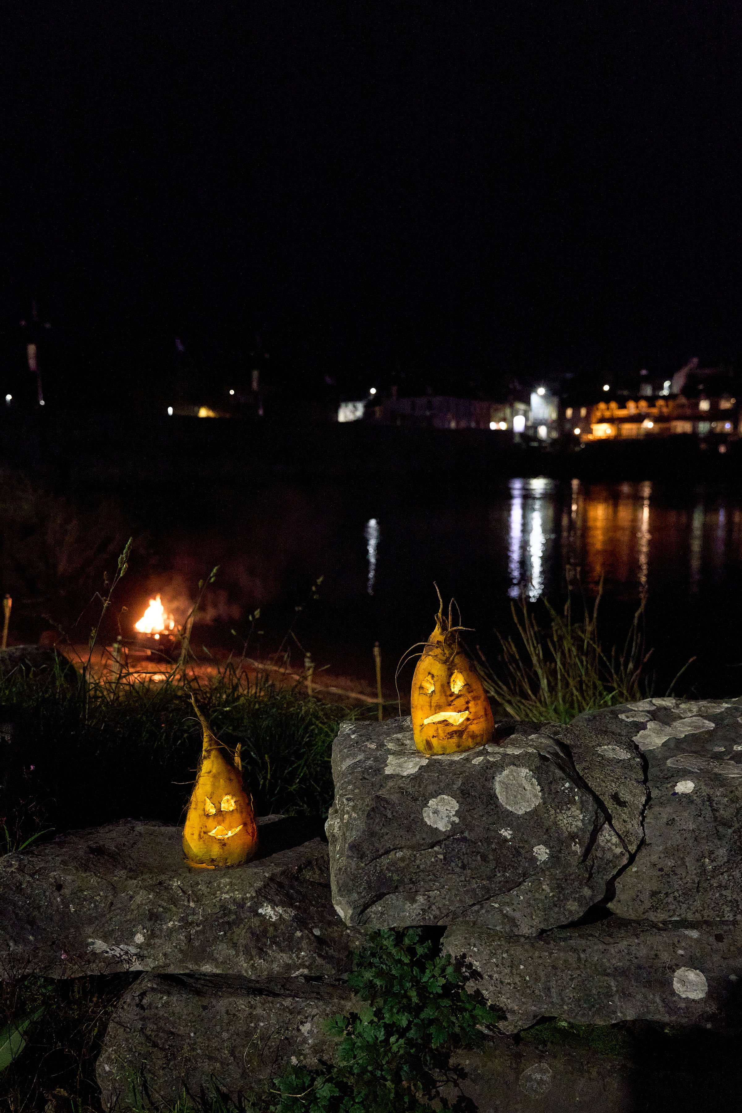 Two carved yellow pumpkins with faces illuminated from inside, sitting on rocks by a waterfront at night, with a small campfire in the background and city lights reflecting on the water.