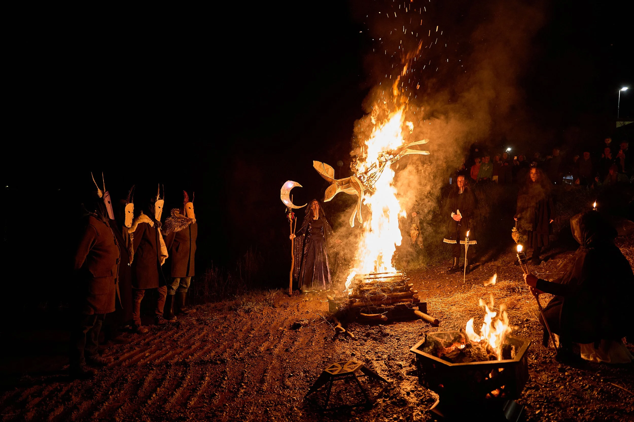 Ancestral Effigy made by Phil McSweeney of Tralee, in the procession on Halloween night.People gathered around a bonfire at night, some wearing masks and costumes with horns and feathers, possibly participating in a traditional ritual or celebration.