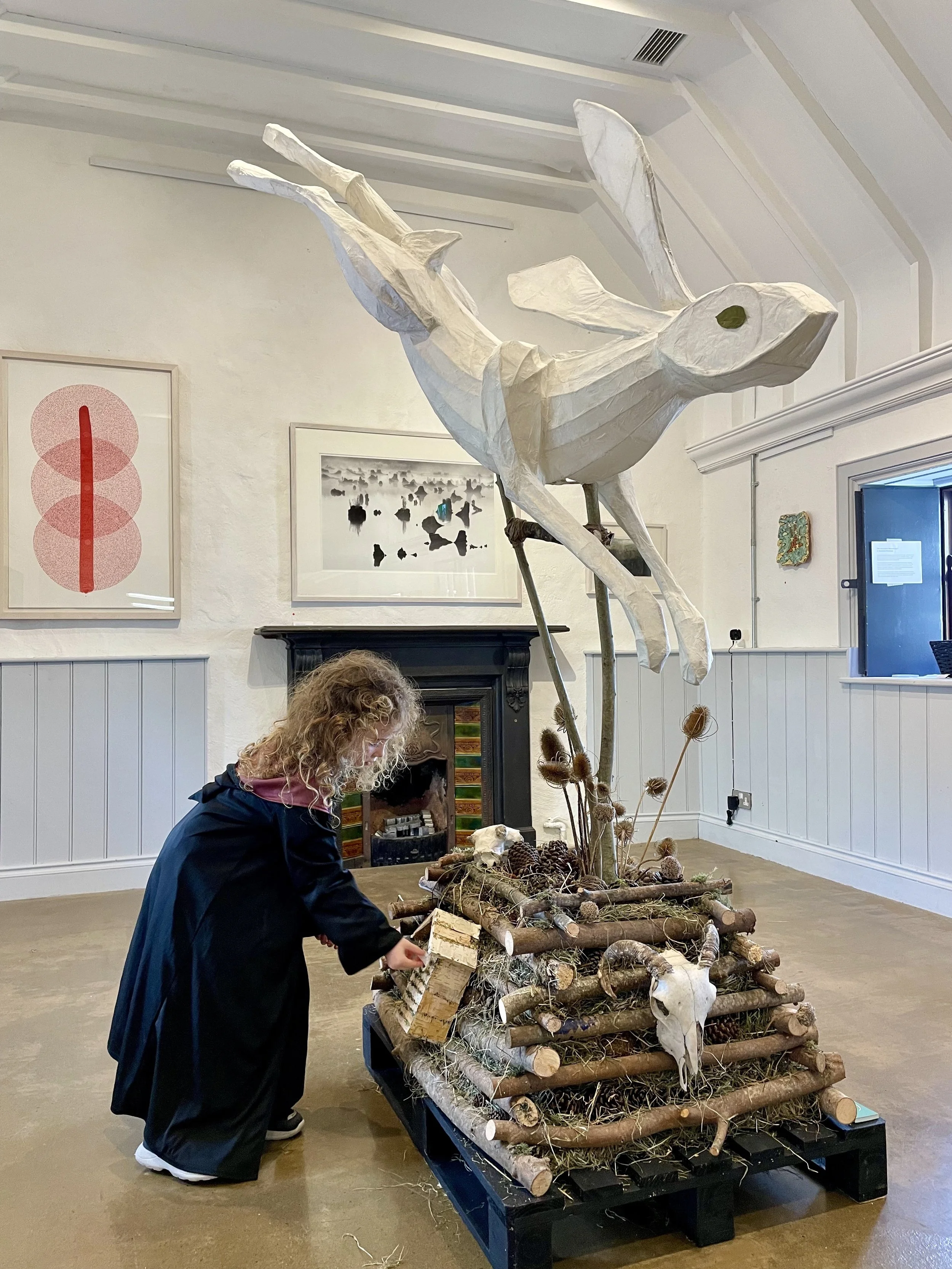 An art installation of a large, white, paper-mâché hare with long legs, mounted on a base decorated with sticks, pine cones, animal skulls, and dried plants. A woman with curly hair wearing a black coat is examining or adjusting the base.