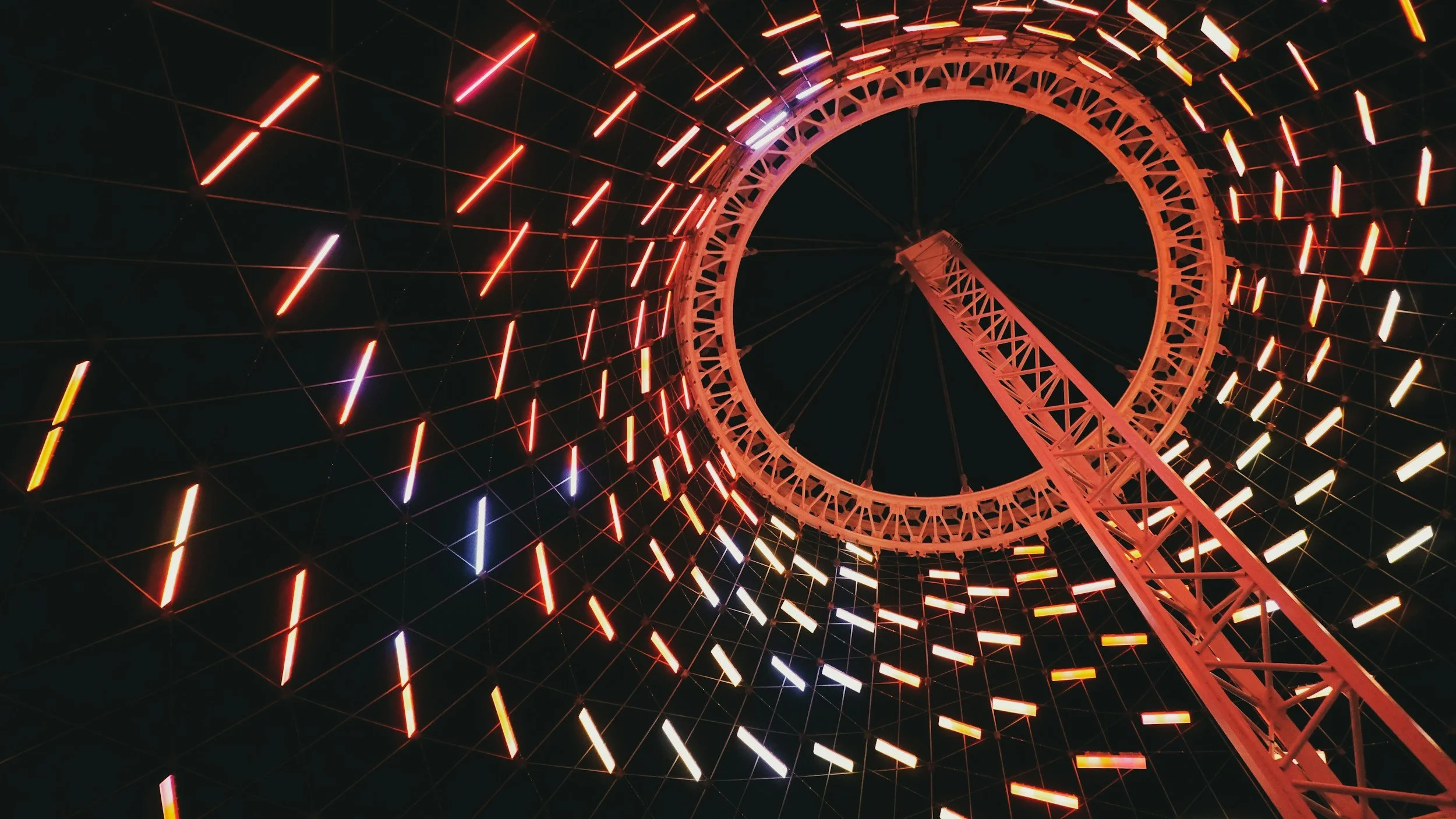 Looking up at a Ferris wheel with colorful lights, viewed from below at night.