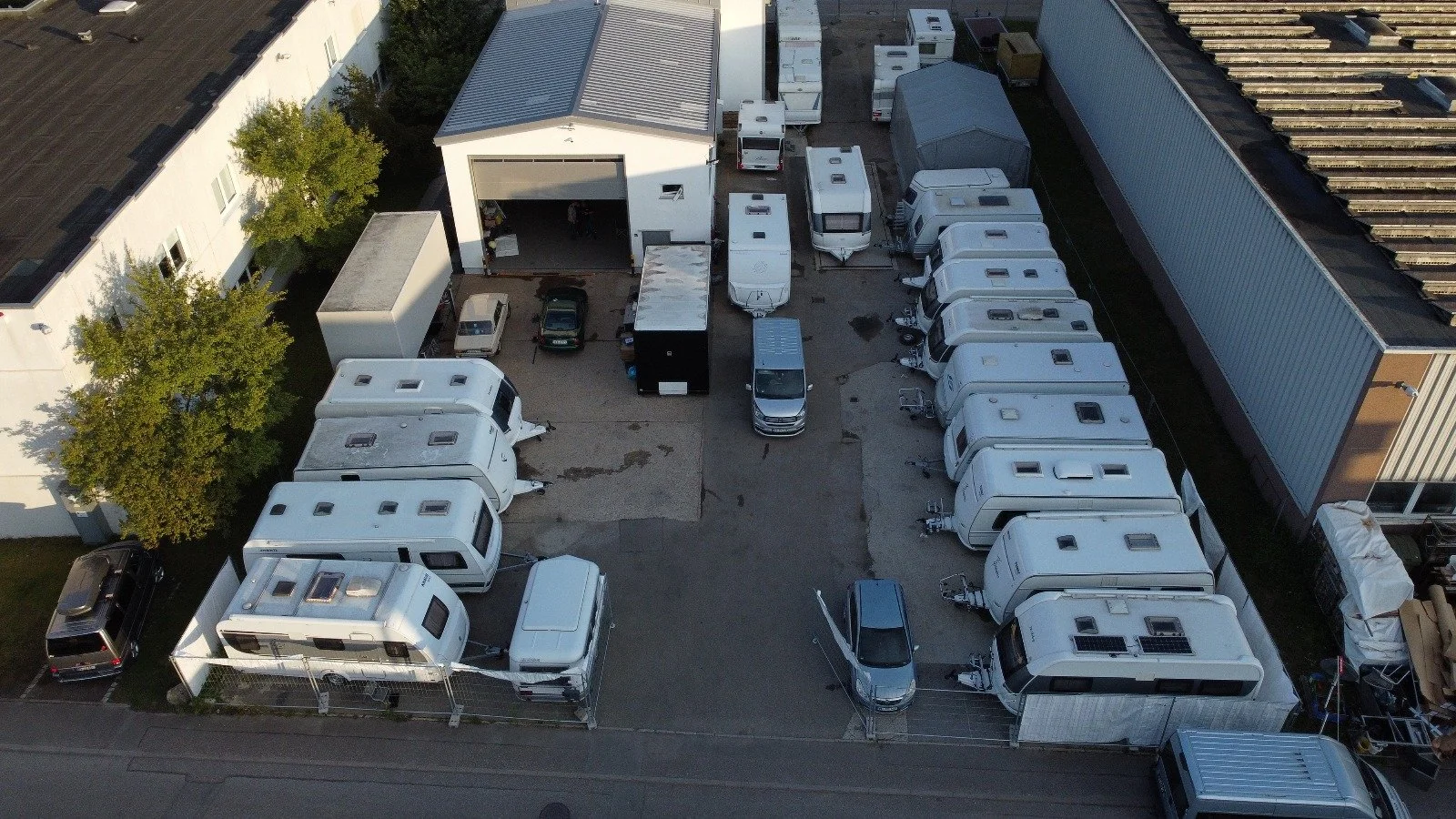 Luftaufnahme eines Parkplatzes mit mehreren Wohnwagen, Fahrzeugen und einer Lagerhalle, umgeben von Gebäuden und Bäumen.