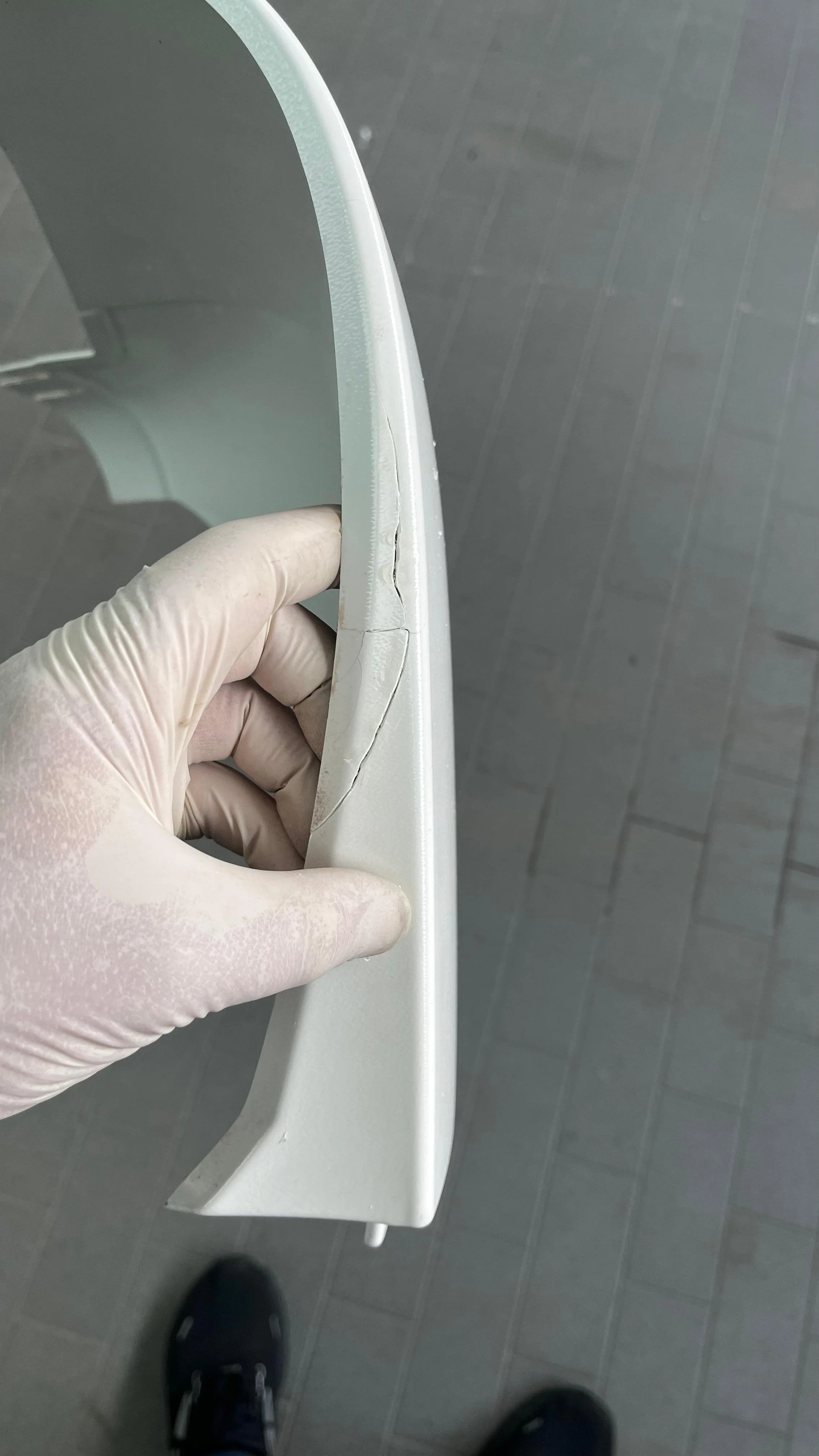 Ein Hand in einem latexfreien Handschuh hält eine zerbrochene weiße Kunststoffverkleidung. Der Boden besteht aus grau gefliesten Fliesen.